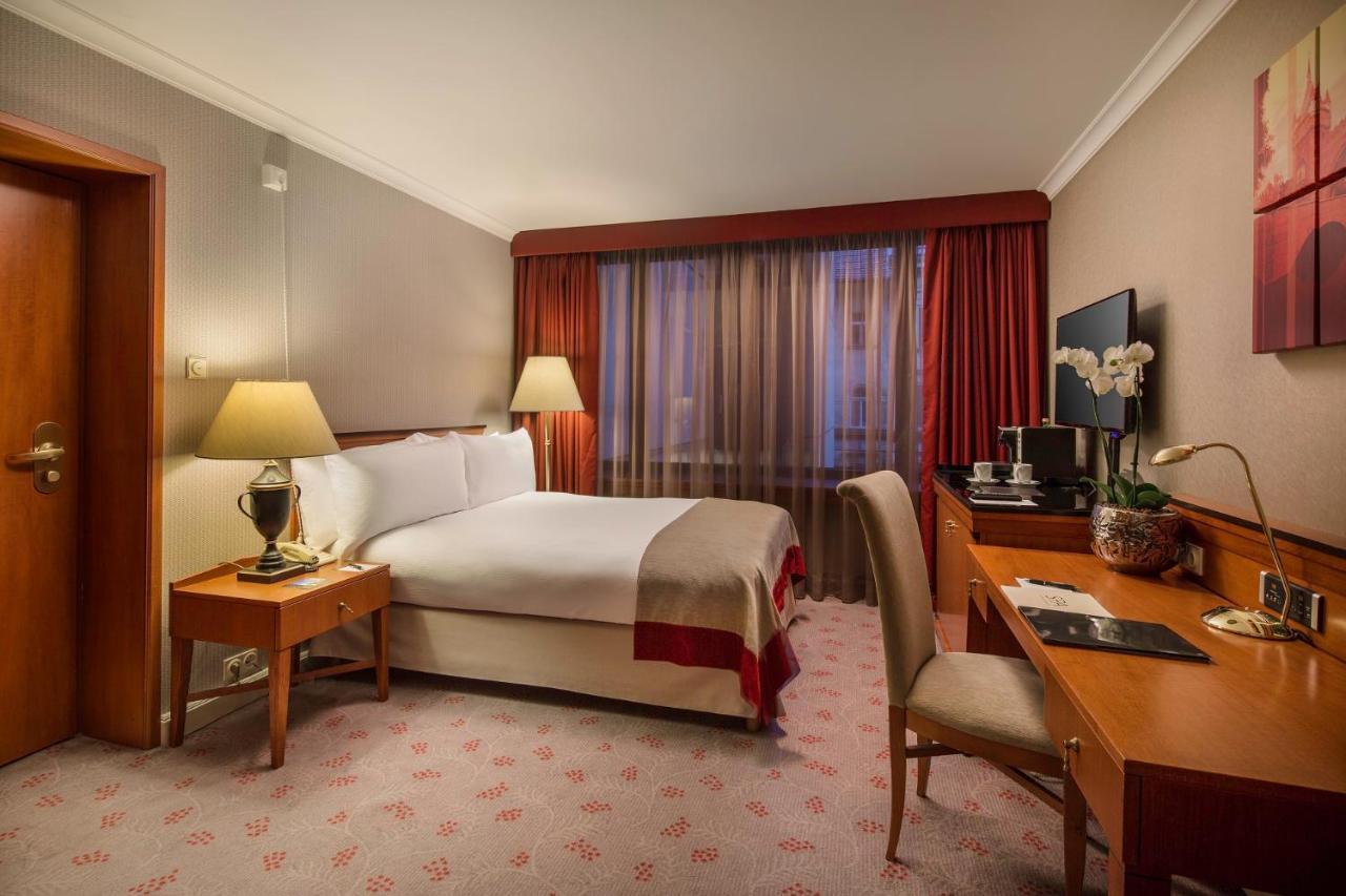 Intercontinental Budapest By Ihg Esterno foto The photo shows a well-furnished hotel room. It features a comfortable double bed with white linens and a decorative throw. There are two bedside tables with lamps on either side of the bed. A large window with sheer curtains lets in natural light, a
