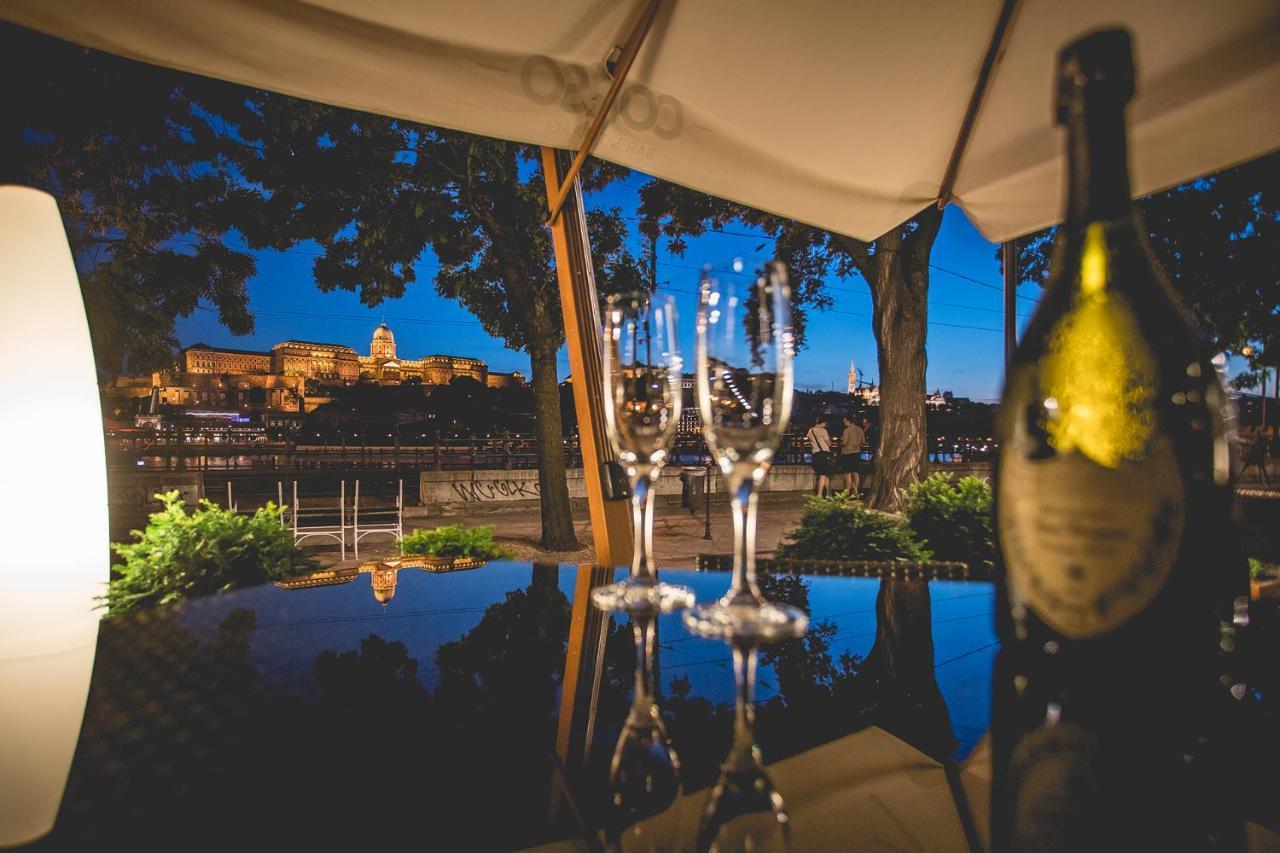 Intercontinental Budapest By Ihg Esterno foto The photo shows a scenic nighttime view of a city landscape, likely by a river, with illuminated buildings in the background. In the foreground, there are two empty champagne flutes placed on a reflective surface, along with an open bottle of champag