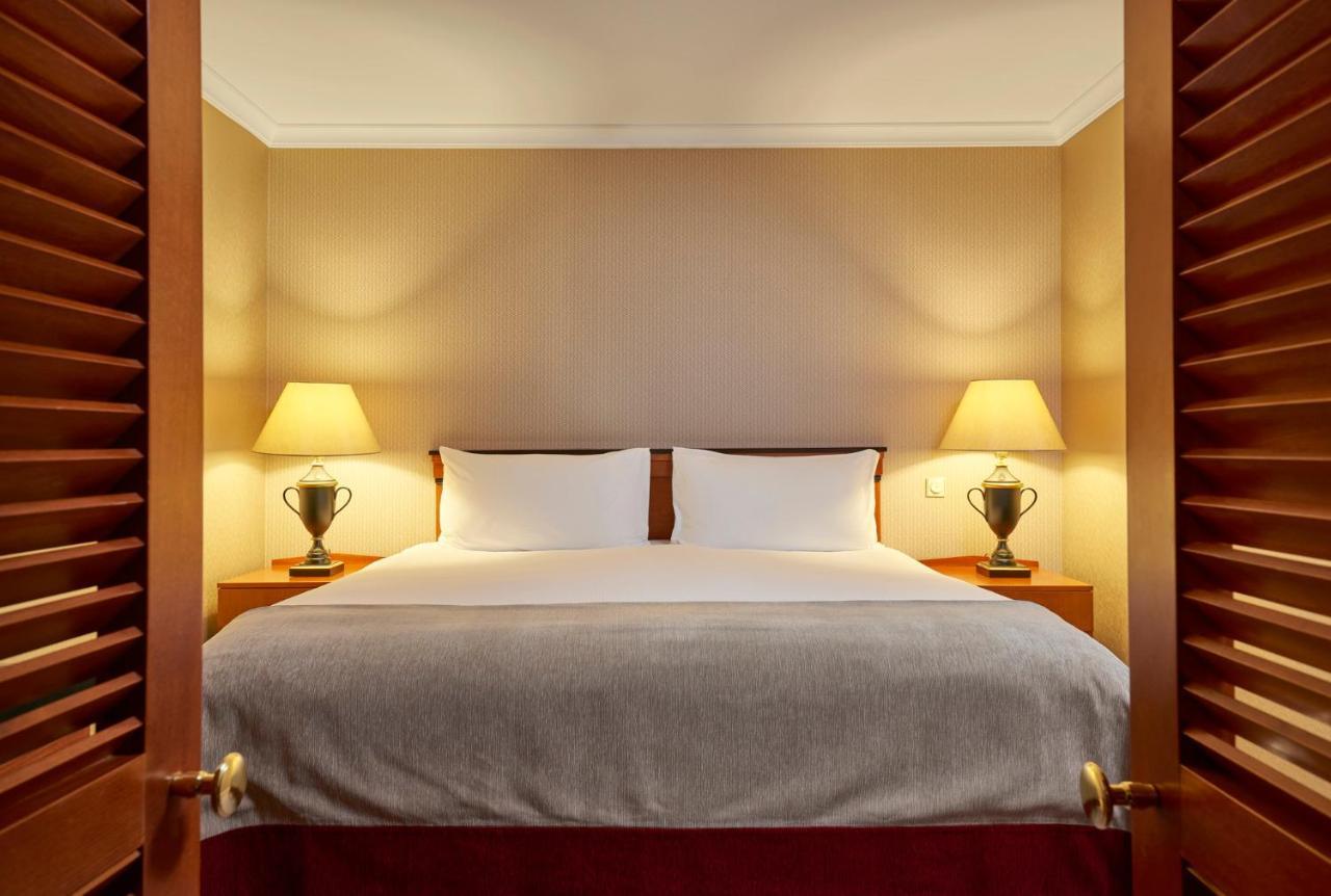 Intercontinental Budapest By Ihg Esterno foto The photo shows a cozy bedroom designed for comfort. In the center, there is a double bed with white linens and a gray blanket at the foot. On either side of the bed, there are matching bedside tables with green lamps providing warm light. The walls 