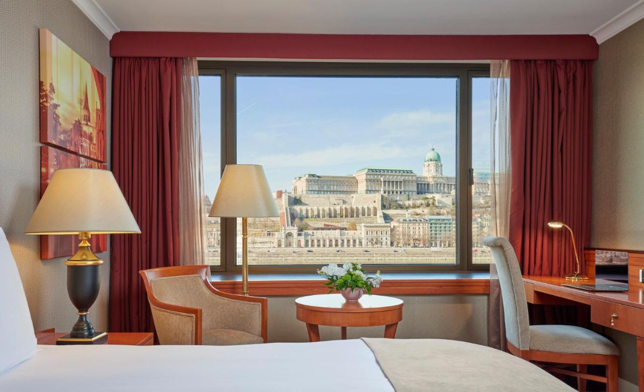 Intercontinental Budapest By Ihg Esterno foto The photo depicts a hotel room with a large window that offers a view of a historic building or castle in the background. The room features elegant decor, including a bed with white linens, a small round table with a floral arrangement, and a couple 