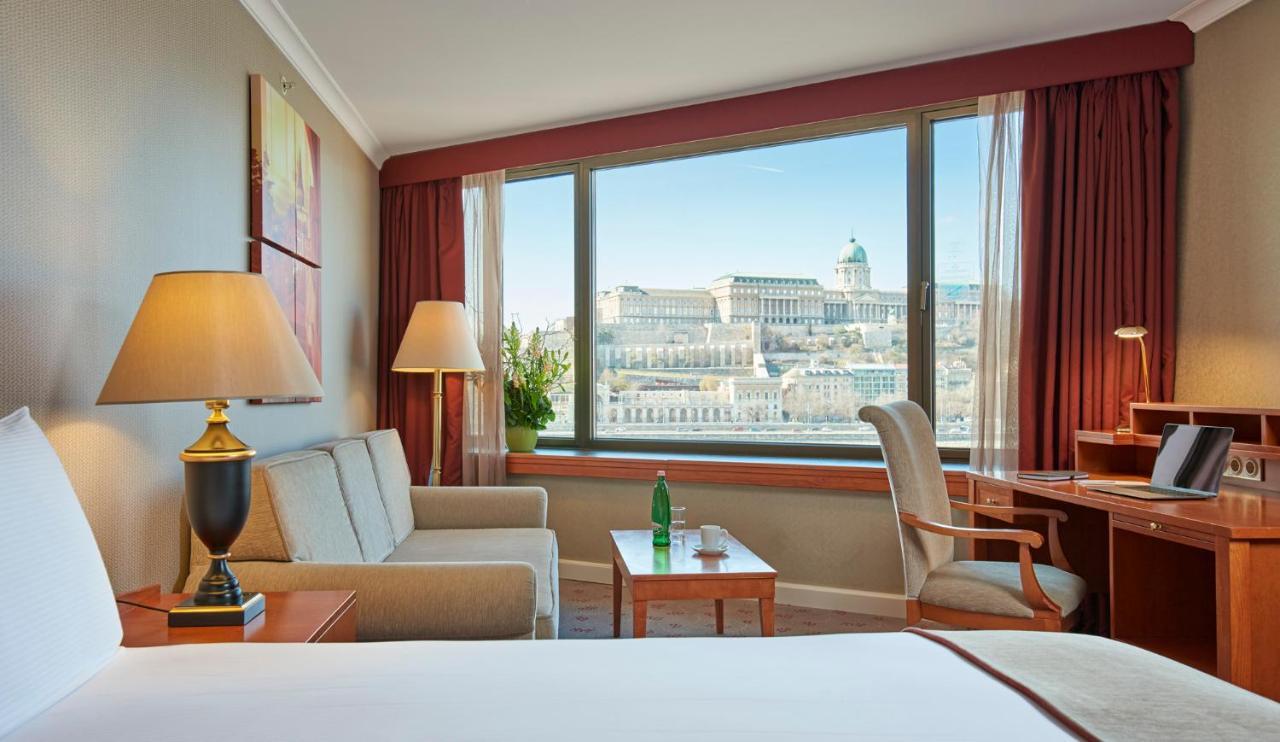 Intercontinental Budapest By Ihg Esterno foto The photo depicts a cozy hotel room featuring a large window with a scenic view. Inside, there are two comfortable chairs and a small table, with a lamp on a side table illuminating the space. A desk is also present, possibly for work. The window off