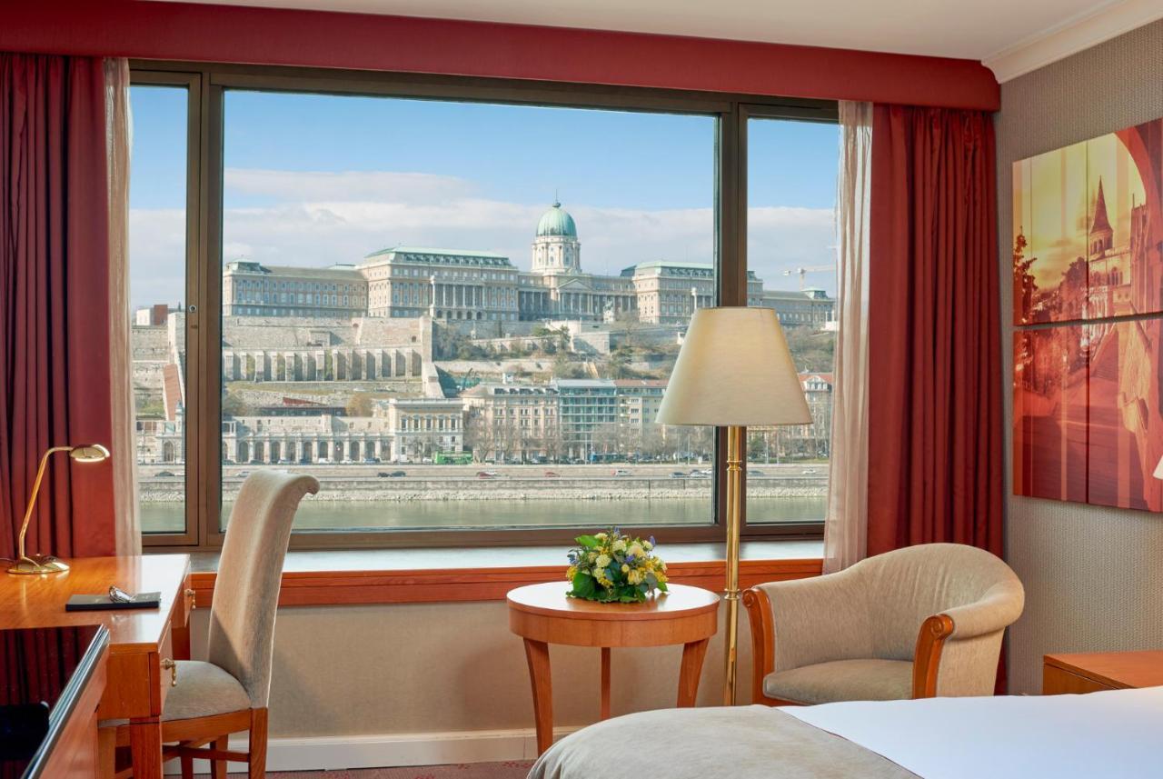 Intercontinental Budapest By Ihg Esterno foto The image shows the interior of a hotel room with a large window that offers a view of a picturesque landscape. Through the window, one can see a historic building situated on a hill, likely part of a castle or palace, and a scenic backdrop featuring