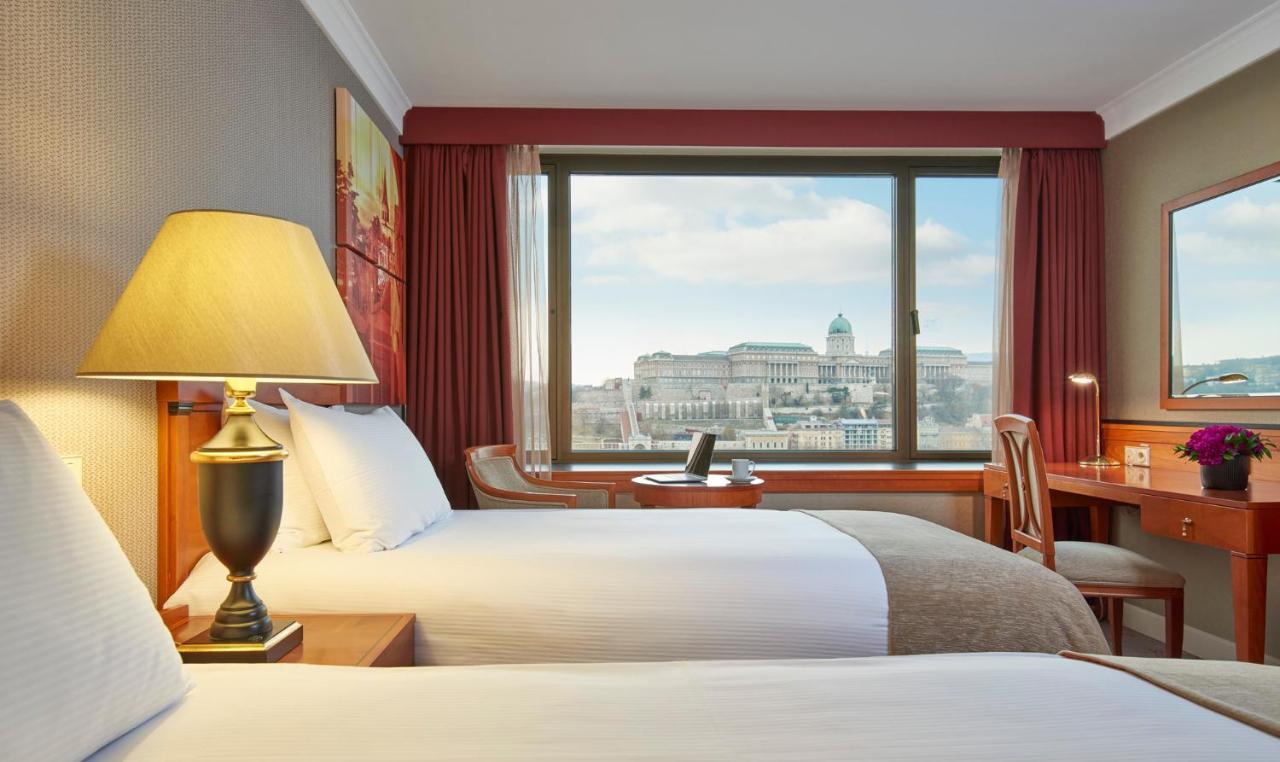 Intercontinental Budapest By Ihg Esterno foto The photo shows a hotel room featuring two beds with white linens and gray accents. There is a lamp on a bedside table, providing soft lighting. A large window offers a view of an outdoor scene, likely showcasing a cityscape with prominent buildings 