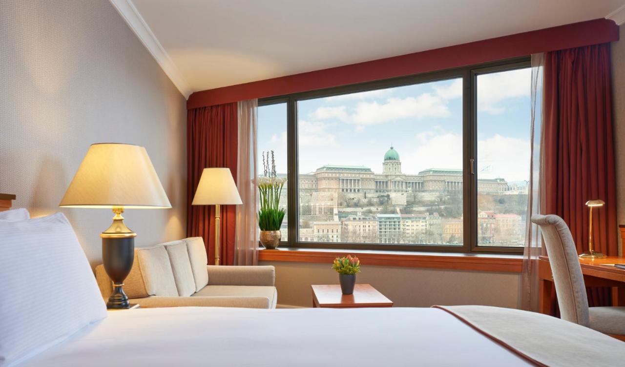 Intercontinental Budapest By Ihg Esterno foto The photo depicts the interior of a hotel room. There is a comfortable seating area with a small table, a lamp, and a window that provides a view of a grand building in the background. The building, likely a historic structure, features a dome and is