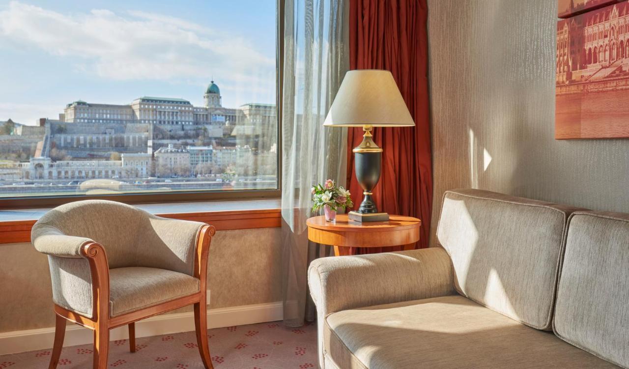 Intercontinental Budapest By Ihg Esterno foto The photo shows a cozy hotel room with a large window offering a view of a cityscape. Inside the room, there is a comfortable-looking beige armchair beside a small round table. On the table, there is a lamp and a small flower arrangement. The walls a