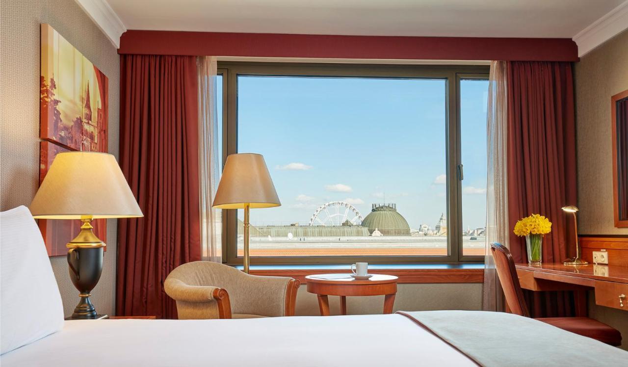 Intercontinental Budapest By Ihg Esterno foto The photo shows the interior of a hotel room with a view through a large window. In the foreground, there is a bed with white bedding, and nearby, a lamp and a small table with a chair. On the table, there's a cup, possibly for tea or coffee. The win