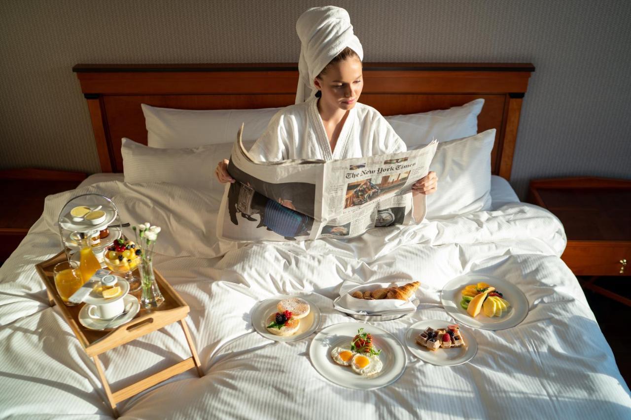 Intercontinental Budapest By Ihg Esterno foto The photo shows a person sitting on a bed, dressed in a white bathrobe with a towel on their head. They are reading a newspaper and surrounded by an assortment of food arranged on plates around them. There are various breakfast items, including pastr