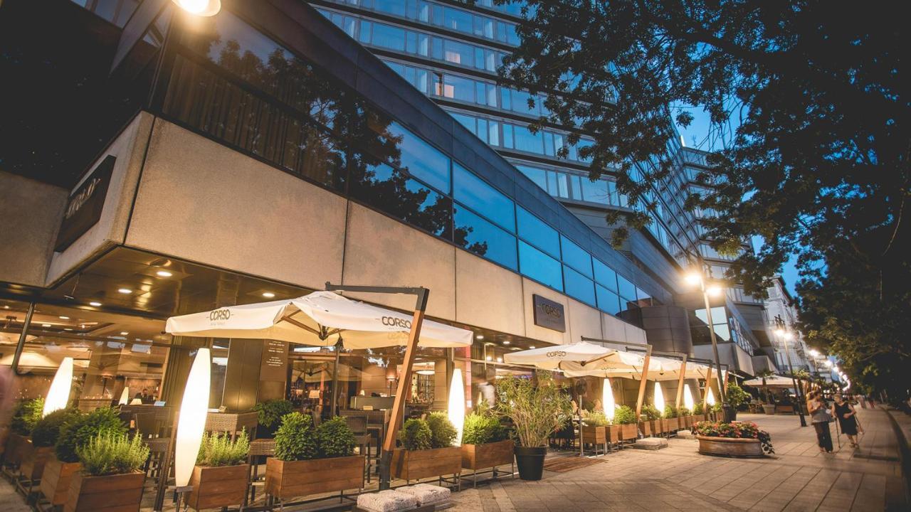 Intercontinental Budapest By Ihg Esterno foto The photo depicts a modern building with large glass windows, likely a restaurant or café, situated on a city street. It features an outdoor seating area with umbrellas providing shade, surrounded by plants and wooden planters. The scene is captured 
