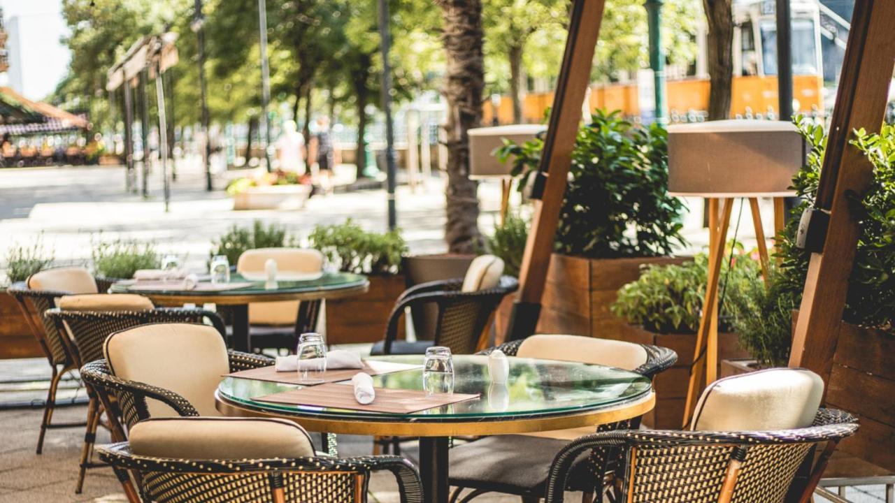 Intercontinental Budapest By Ihg Esterno foto The photo shows an outdoor dining area of a restaurant. There are several round tables set with chairs, some of which have cushions. The tables feature glass tops, and condiments or glassware can be seen on them. Surrounding the seating area are gree