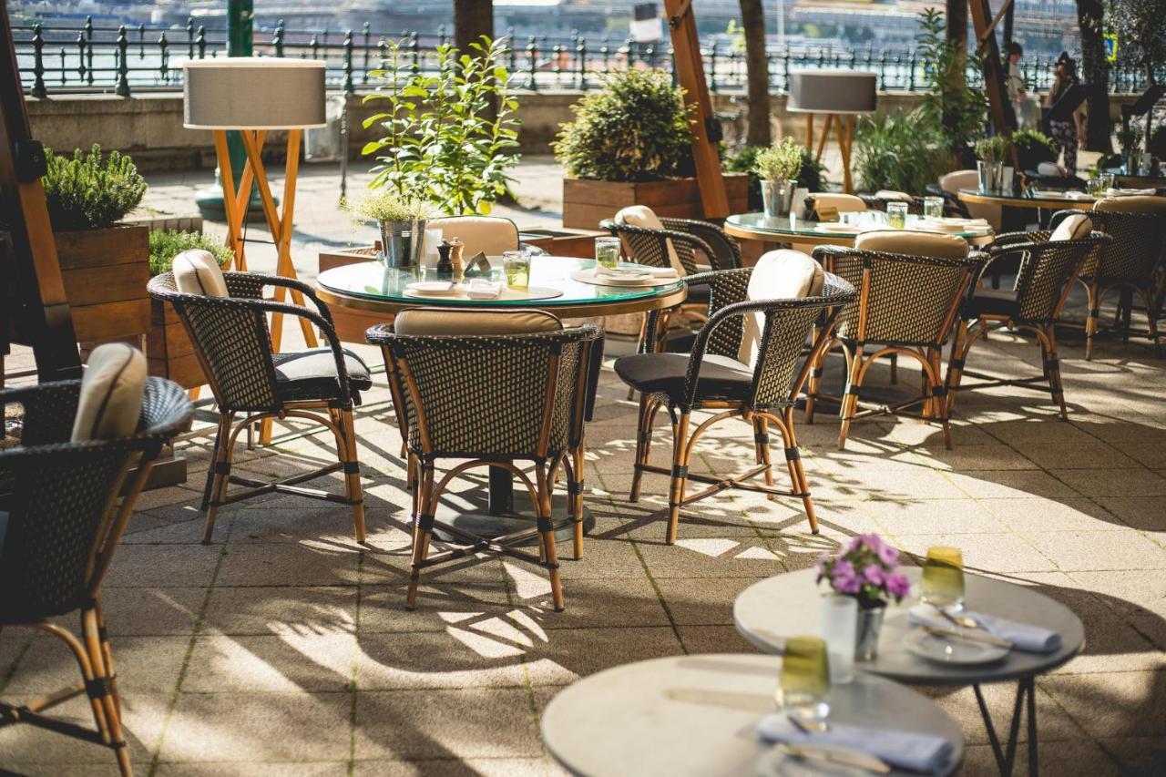 Intercontinental Budapest By Ihg Esterno foto The photo depicts an outdoor dining area featuring several round tables and chairs arranged on a patio. The tables are decorated with small flower arrangements and have place settings, suggesting a ready-to-dine setup. There are potted plants surroun