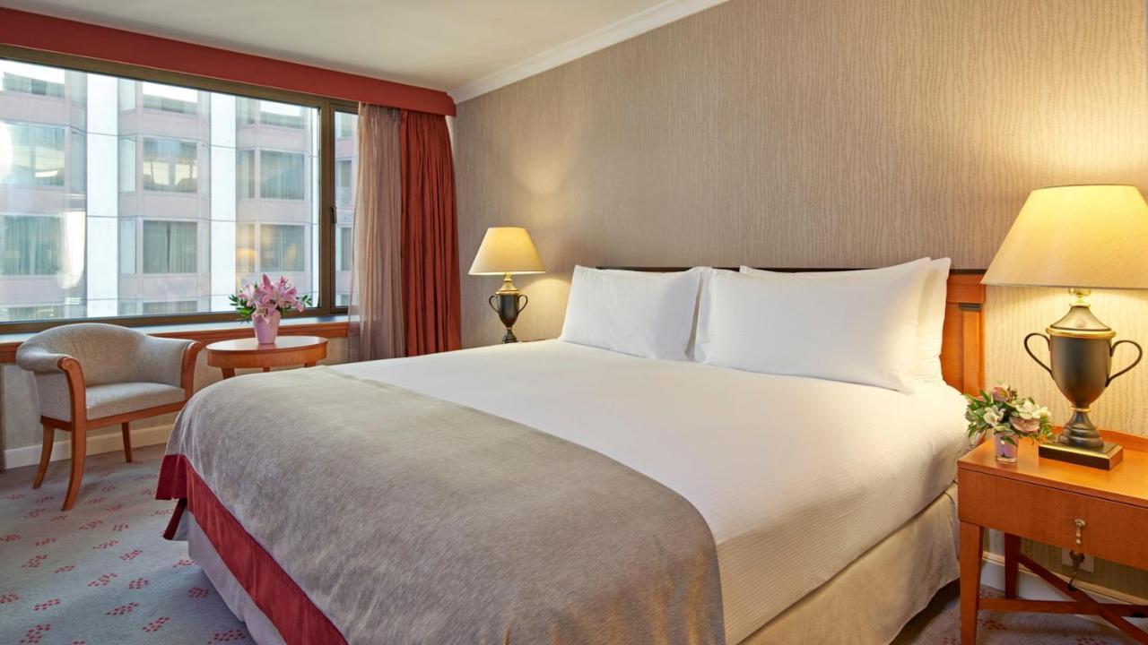 Intercontinental Budapest By Ihg Esterno foto The photo shows a hotel room with a large bed featuring white bedding and two pillows. The walls are light-colored with a subtle pattern, and there are large windows allowing natural light to enter. Next to the bed, there are bedside lamps providing 
