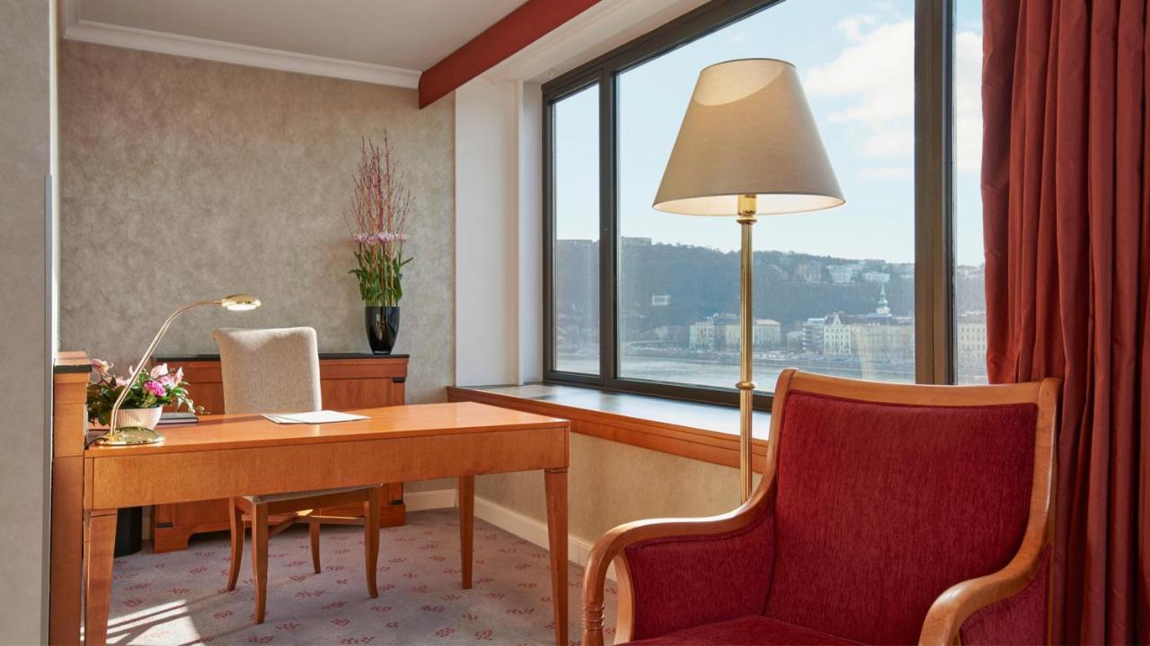 Intercontinental Budapest By Ihg Esterno foto The photo shows a stylish room, likely an office or a hotel suite. There's a wooden desk placed near a large window with a view of the outside landscape. A comfortable red armchair sits next to the desk, providing a cozy seating option. The room feat