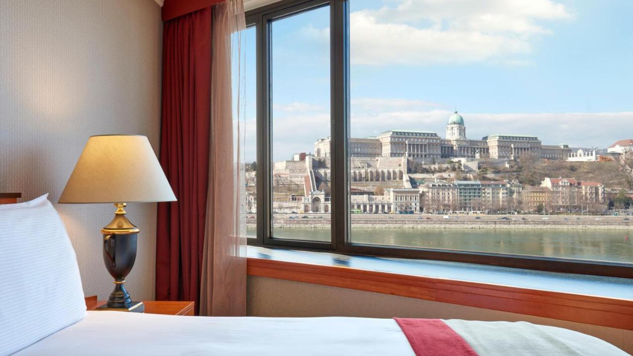 Intercontinental Budapest By Ihg Esterno foto The photo shows a view from a hotel room with a window overlooking a river and a historic building on a hillside. In the foreground, there is a bedside lamp, and you can see the soft furnishings in the room, such as a curtain and part of a bed. The l