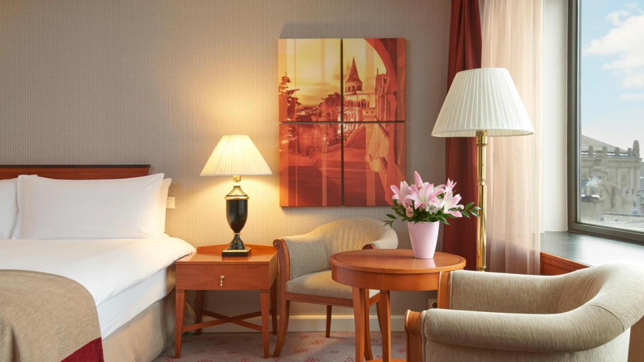 Intercontinental Budapest By Ihg Esterno foto The image shows a cozy hotel room featuring a bed with white linens, a small wooden table with a vase of pink flowers, and a comfortable chair. There's a decorative lamp on the table and another lamp next to a seating area. A piece of wall art depict