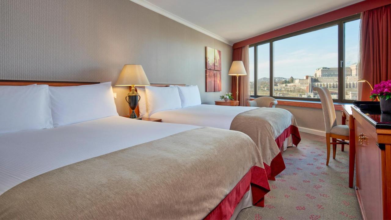 Intercontinental Budapest By Ihg Esterno foto The photo shows a hotel room featuring two twin beds with soft, light-colored bedding. There are bedside lamps on each side of the beds, and a decorative piece of artwork hangs on the wall. A window provides natural light and offers a view of the out