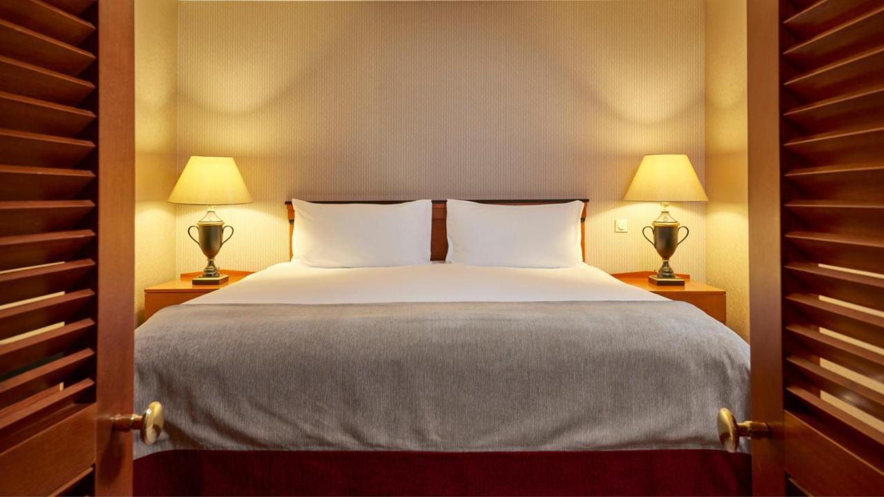 Intercontinental Budapest By Ihg Esterno foto The photo shows a cozy hotel bedroom with a large double bed centered in the frame. The bed has a neatly made white duvet and two white pillows. On either side of the bed, there are bedside tables with table lamps, adding a warm glow to the room. The