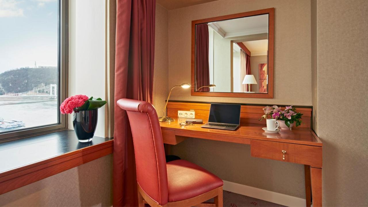 Intercontinental Budapest By Ihg Esterno foto The photo shows a small workspace set up in a room. It features a wooden desk with a laptop on it and a lamp for lighting. There is a comfortable red chair positioned at the desk. To the left of the desk, there is a window with a view, partially cove