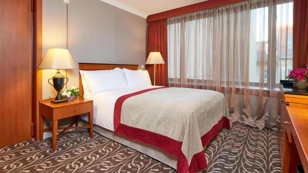 Intercontinental Budapest By Ihg Esterno foto The photo shows a hotel room featuring a neatly made bed with white linens and a light-colored blanket accented with a red border. There is a nightstand next to the bed with a decorative lamp and a small plant. A large window with sheer curtains allo
