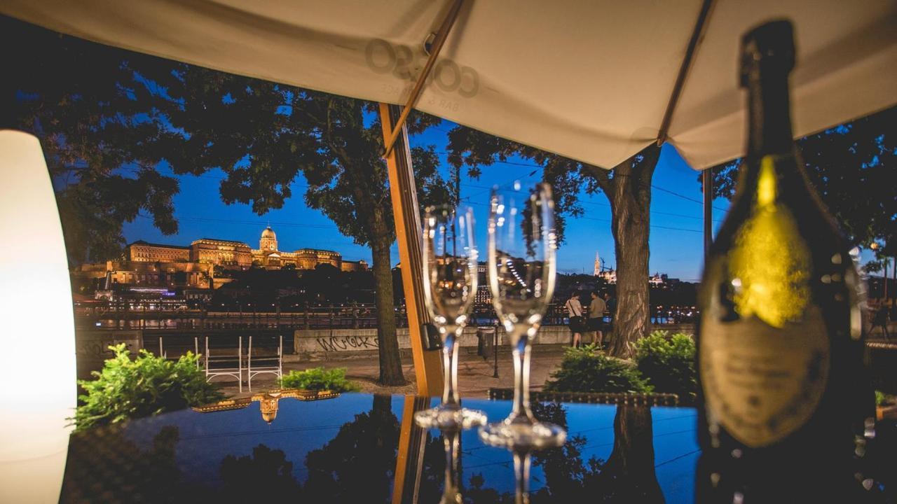 Intercontinental Budapest By Ihg Esterno foto The photo shows a beautiful evening scene featuring a few elegant glasses placed on a reflective surface. In the background, there is a stunning view of a lit-up cityscape, likely featuring a castle or historical building illuminated at night. The at