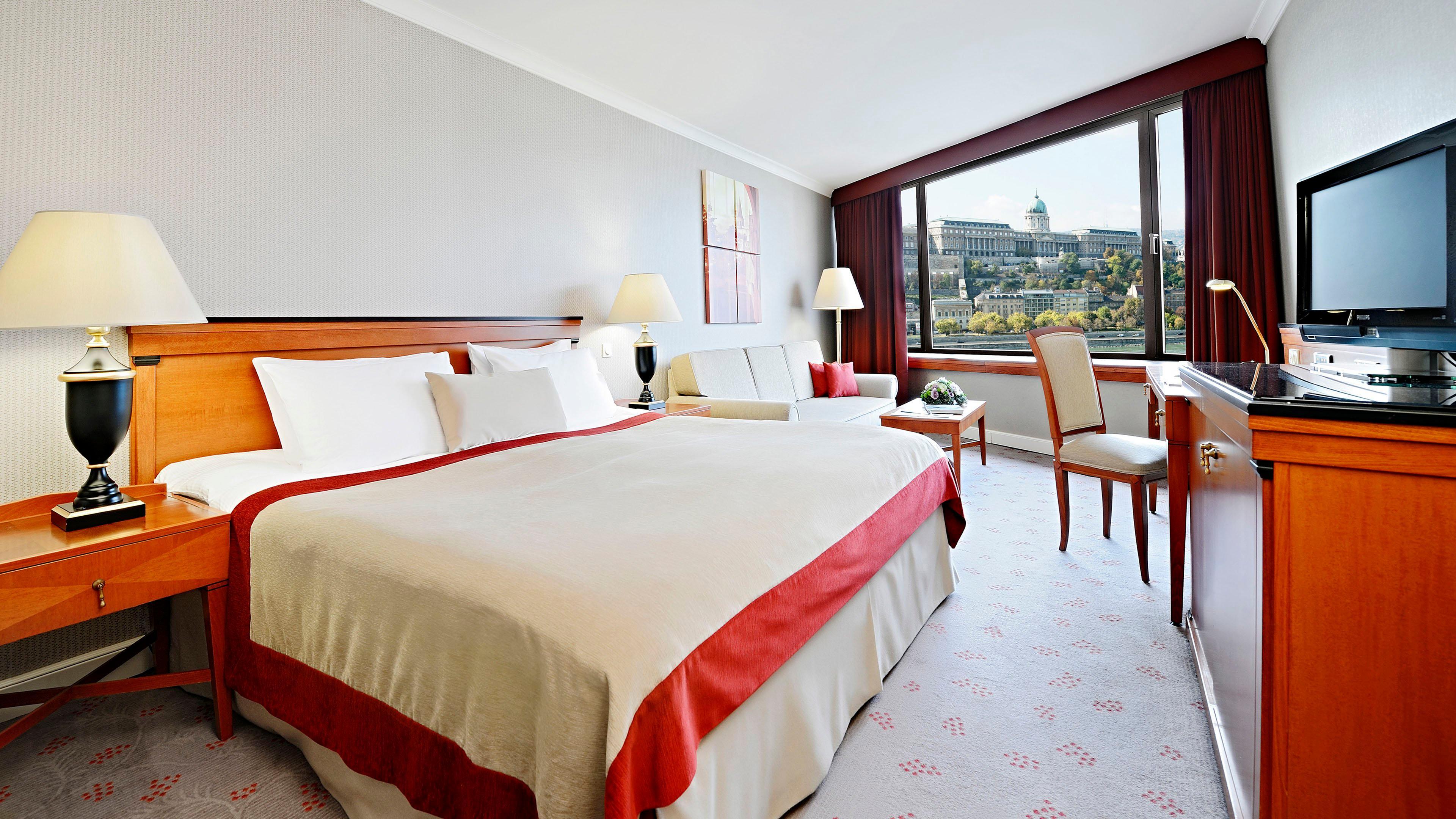 Intercontinental Budapest By Ihg Esterno foto The photo shows a hotel room featuring a large bed with a cream-colored bedspread that has a red border. There are two bedside lamps on either side of the bed, and a comfortable-looking couch is positioned near a window. The room has a desk with a ch