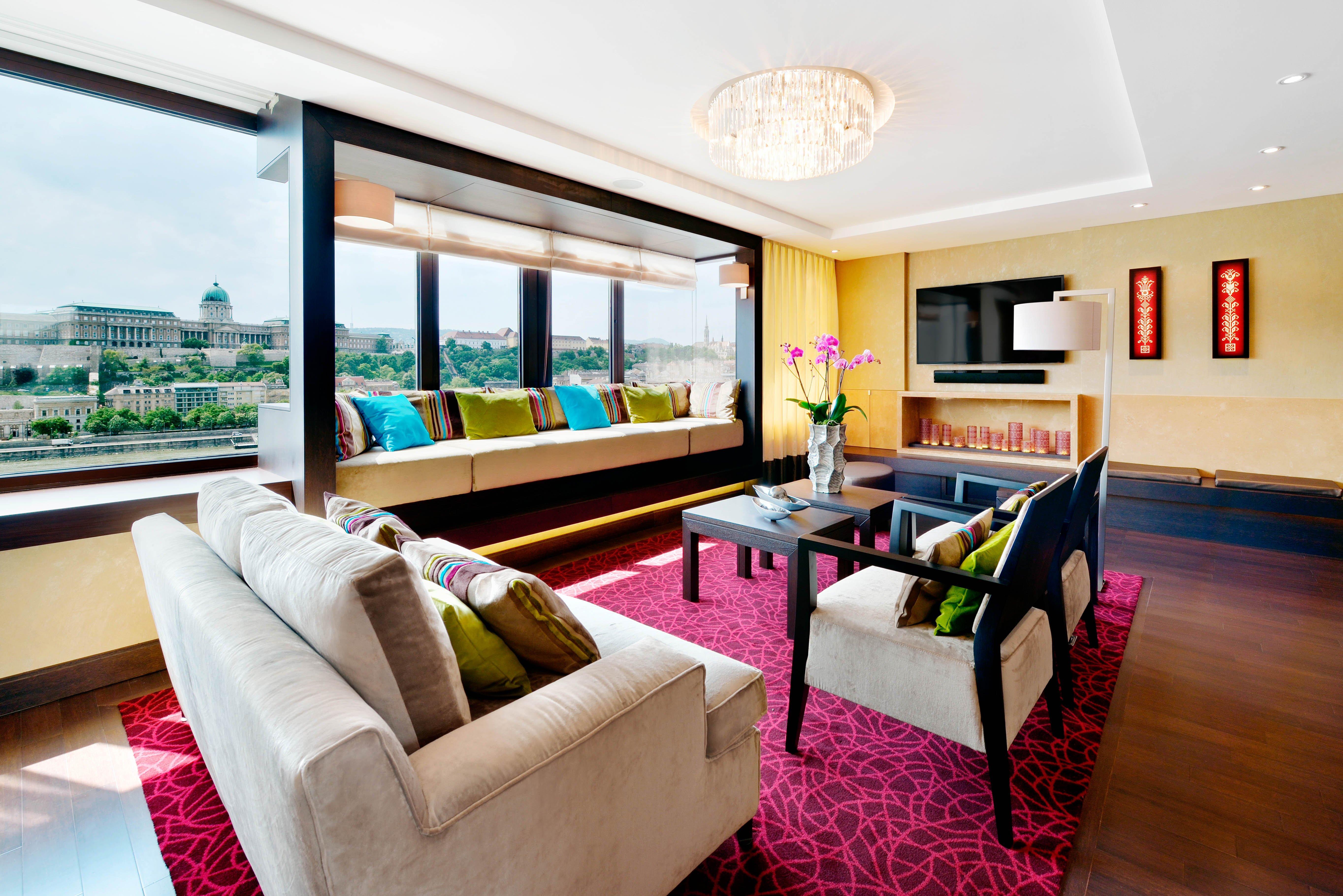 Intercontinental Budapest By Ihg Esterno foto The photo shows a modern and stylish living room with large windows that provide a view of the outside. The space features a neutral-colored sofa adorned with colorful pillows and a matching armchair. There is a dark wooden coffee table in the center