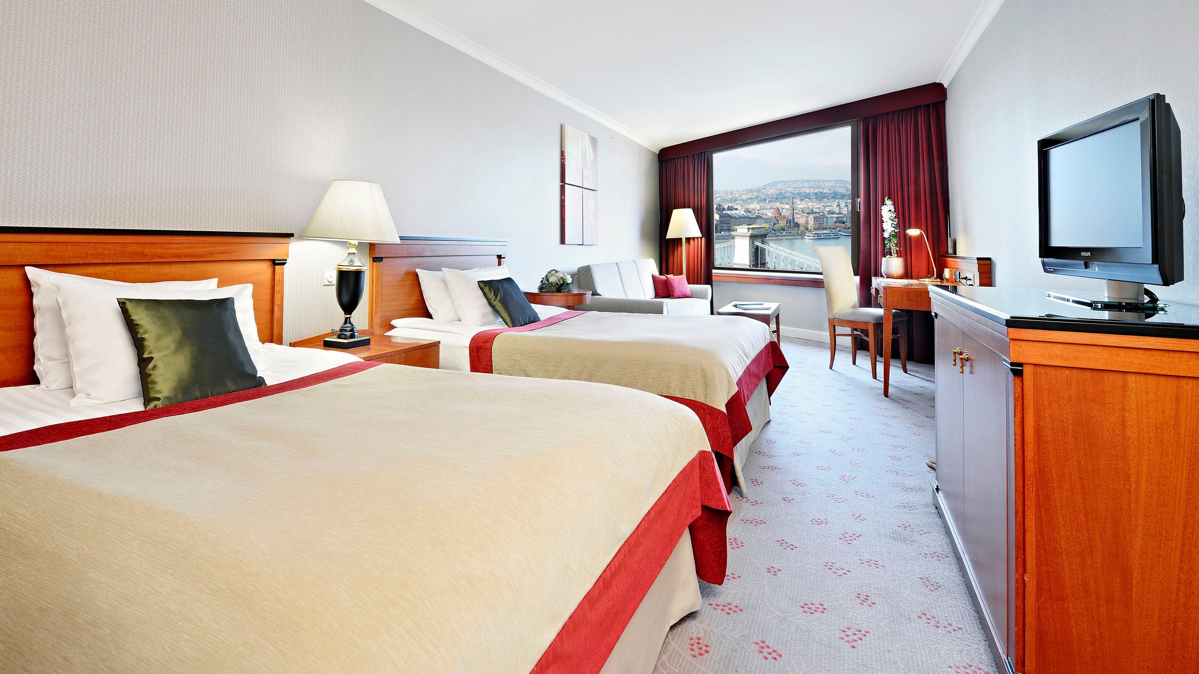 Intercontinental Budapest By Ihg Esterno foto The photo shows a hotel room featuring two double beds covered with light-colored bedding that has a red accent. There are bedside lamps on each side of the beds. A window with a view can be seen, and the room has a warm, inviting color scheme with n