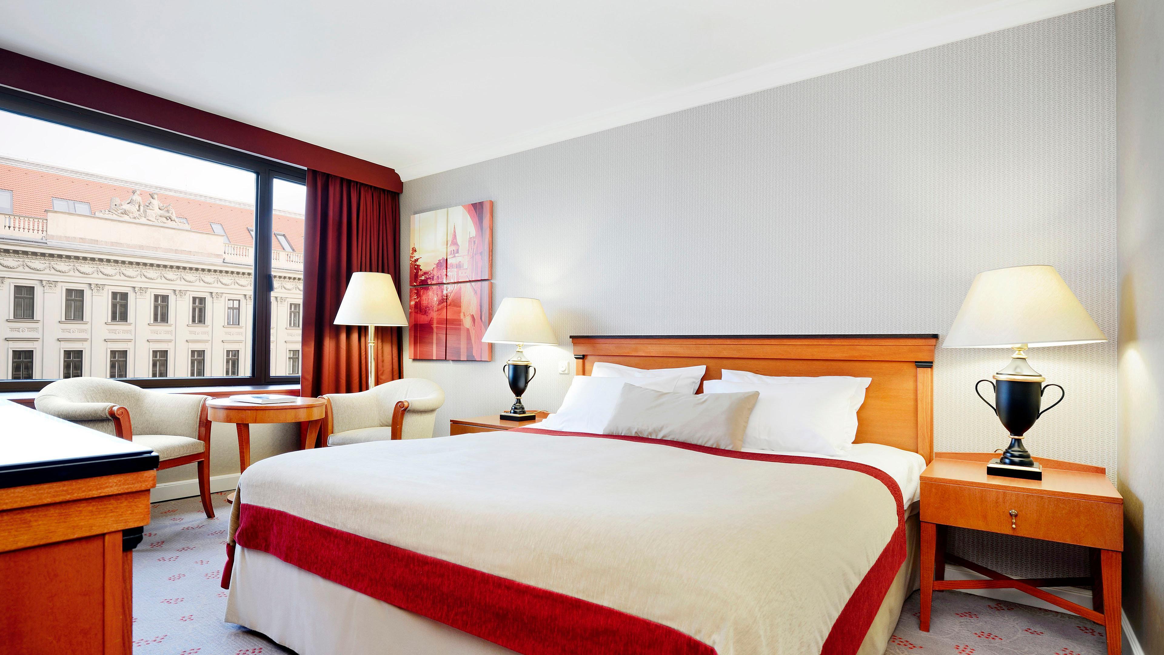 Intercontinental Budapest By Ihg Esterno foto The photo shows a hotel room featuring a large bed with white linens and a decorative red throw. There is a window with a red curtain, allowing natural light to enter the space. Next to the bed, there are two bedside tables with table lamps. A chair 
