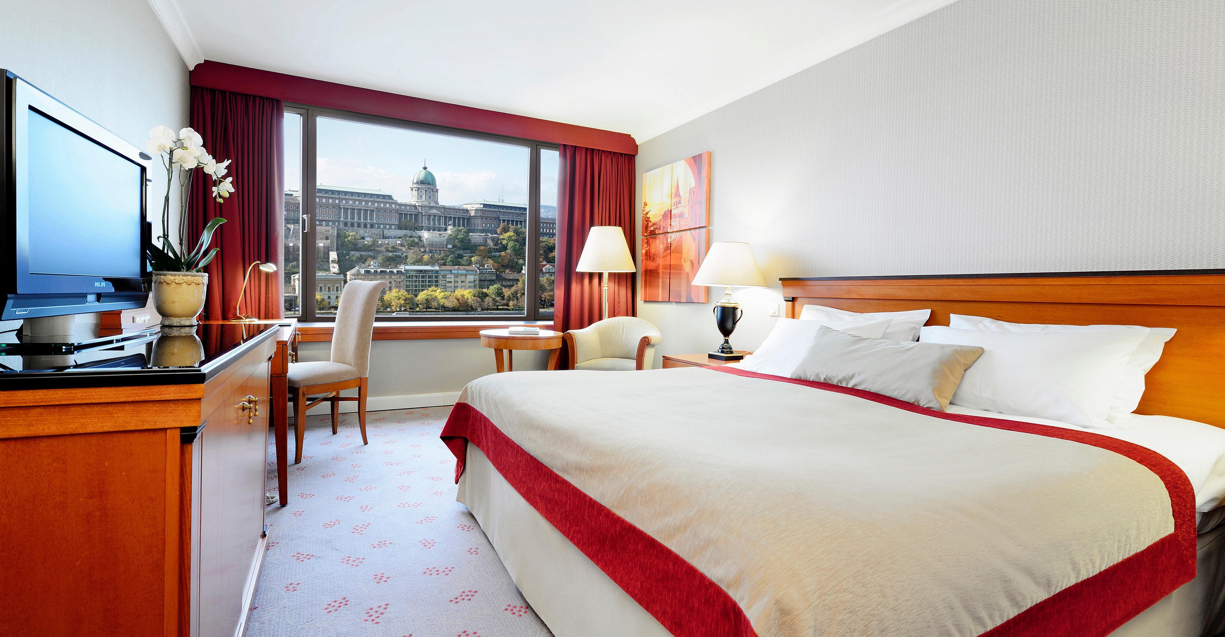 Intercontinental Budapest By Ihg Esterno foto The photo shows a hotel room featuring a large bed with white linens and a red bedspread. There is a wooden desk and chair positioned next to a window that offers a view of a building or architectural structure outside. The room is decorated with sof