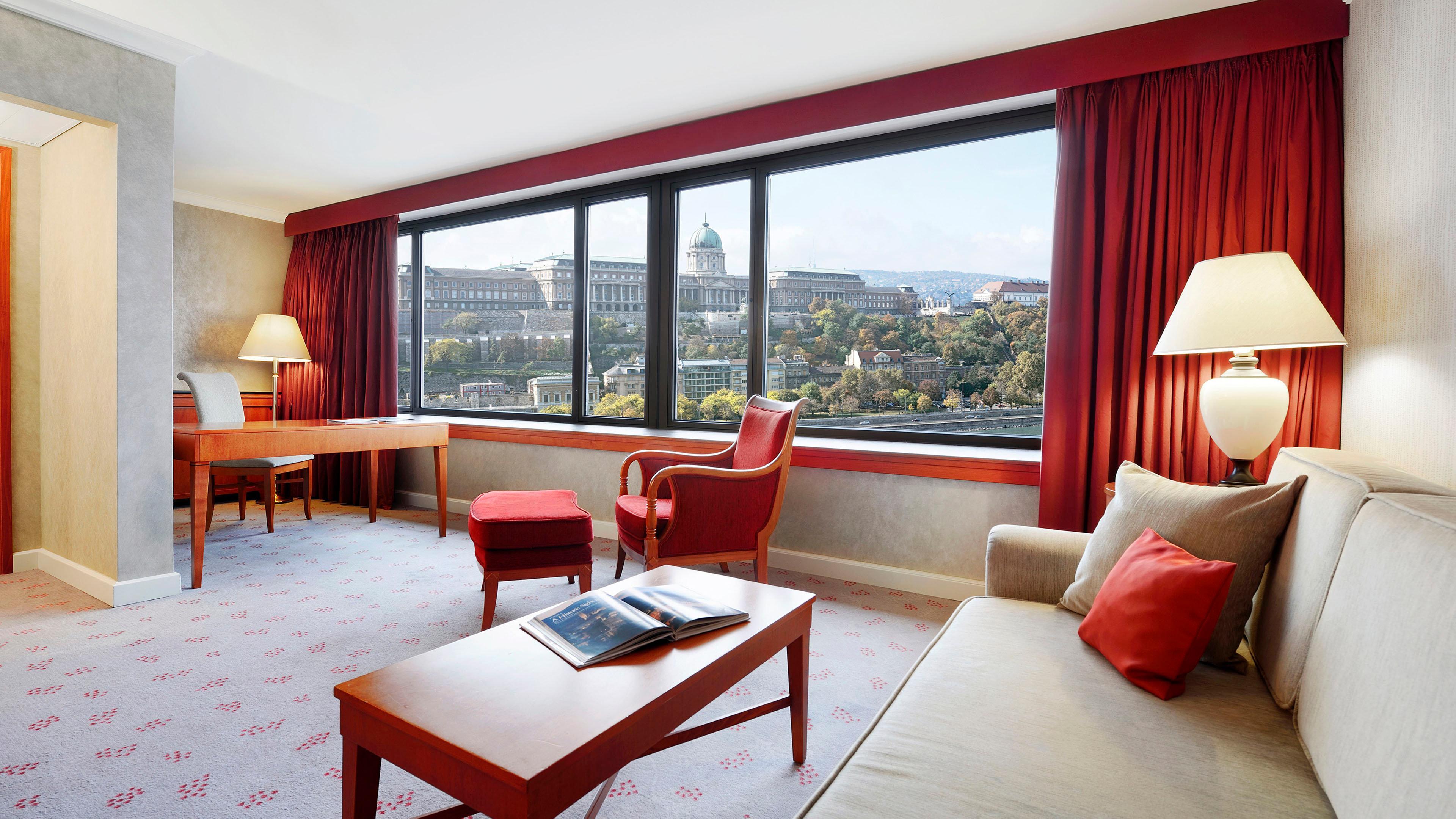 Intercontinental Budapest By Ihg Esterno foto The photo shows a hotel room with a modern and comfortable design. It features a large window that provides a view of a cityscape, including historic buildings and lush trees. Inside the room, there are two armchairs, a small table, and a desk with a