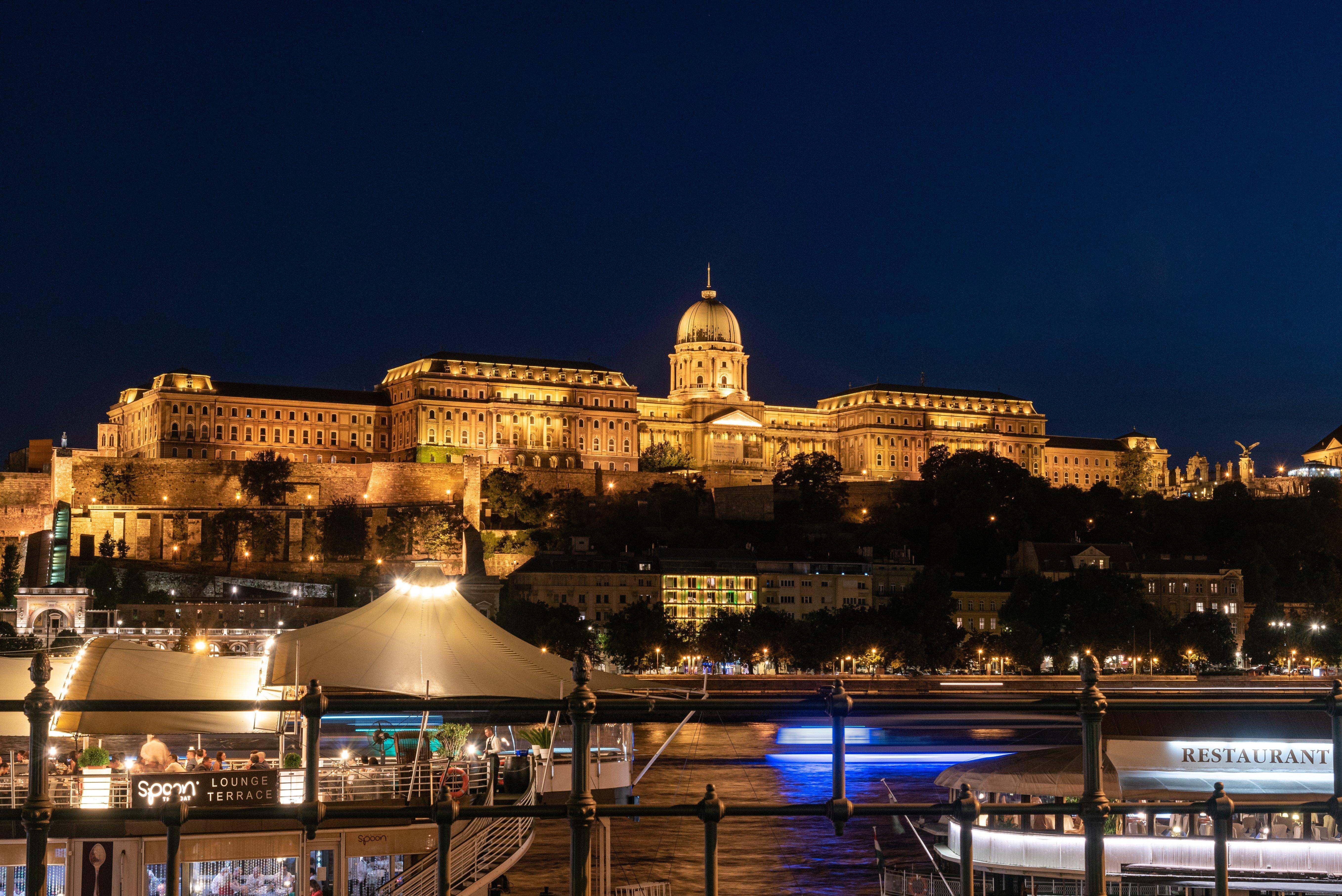 Intercontinental Budapest By Ihg Esterno foto The photo shows a stunning nighttime view of a large, illuminated building complex situated on a hill. The architecture appears grand, with a prominent dome on top and a series of arches and windows. Below the hill, there is a river with boats visibl