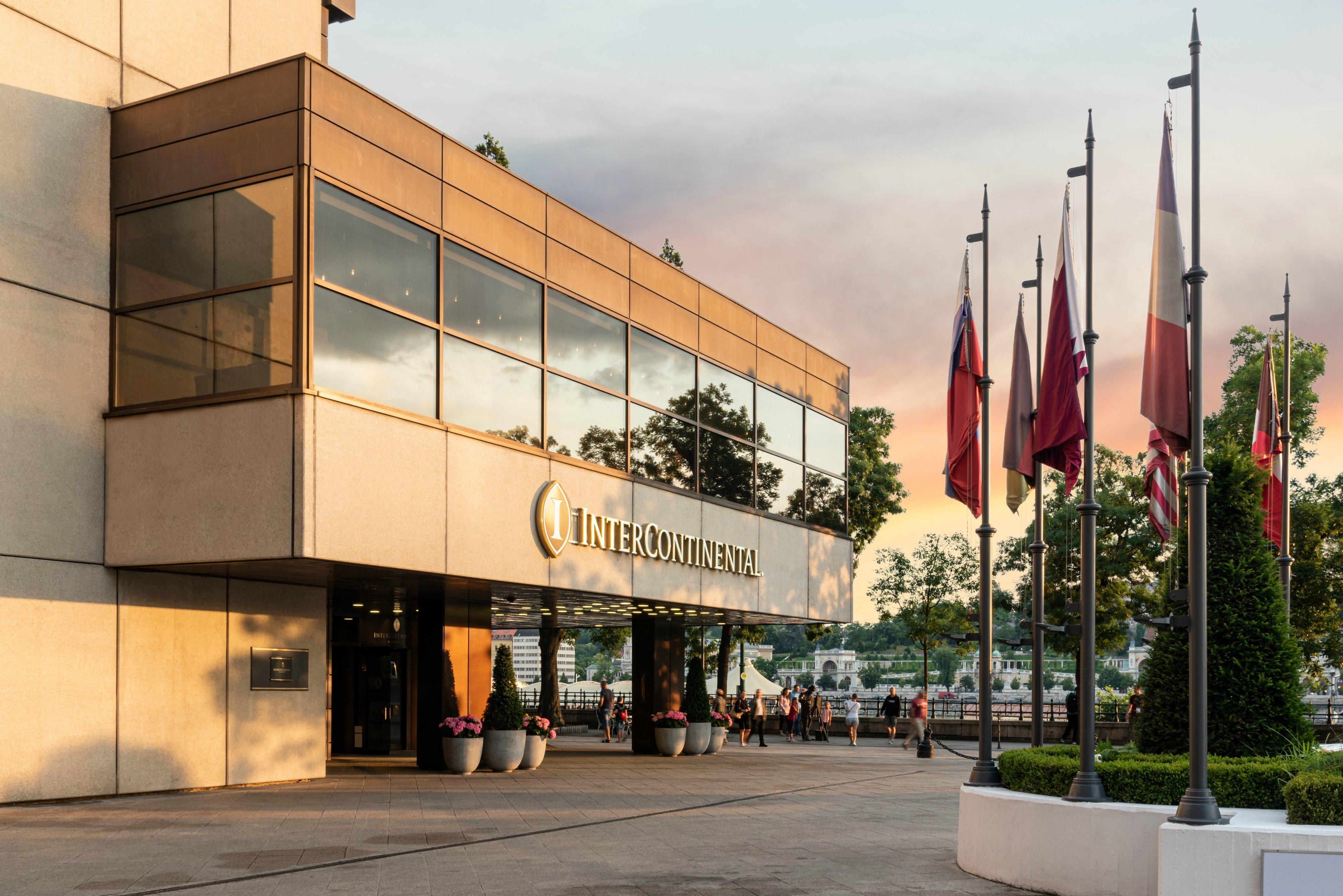 Intercontinental Budapest By Ihg Esterno foto The photo shows the exterior of a building, identified as the Intercontinental Hotel. It features a modern design with large glass windows and a warm color palette. In front of the building, there are several flagpoles displaying various flags. The s