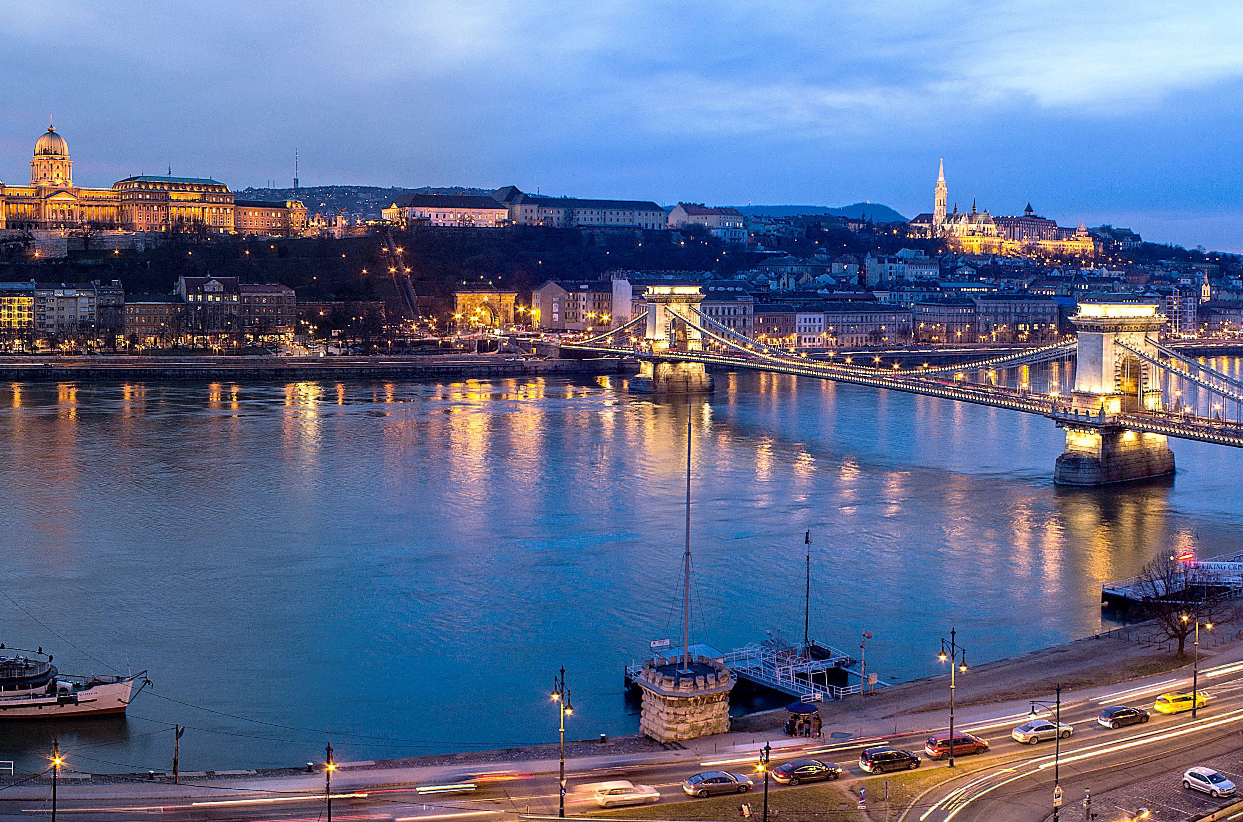 Intercontinental Budapest By Ihg Esterno foto The photo depicts a scenic view of a city, likely during twilight. In the foreground, there is a river that reflects the soft glow of lights from the surrounding structures. A prominent bridge spans the river, connecting two sides of the city. 

On o