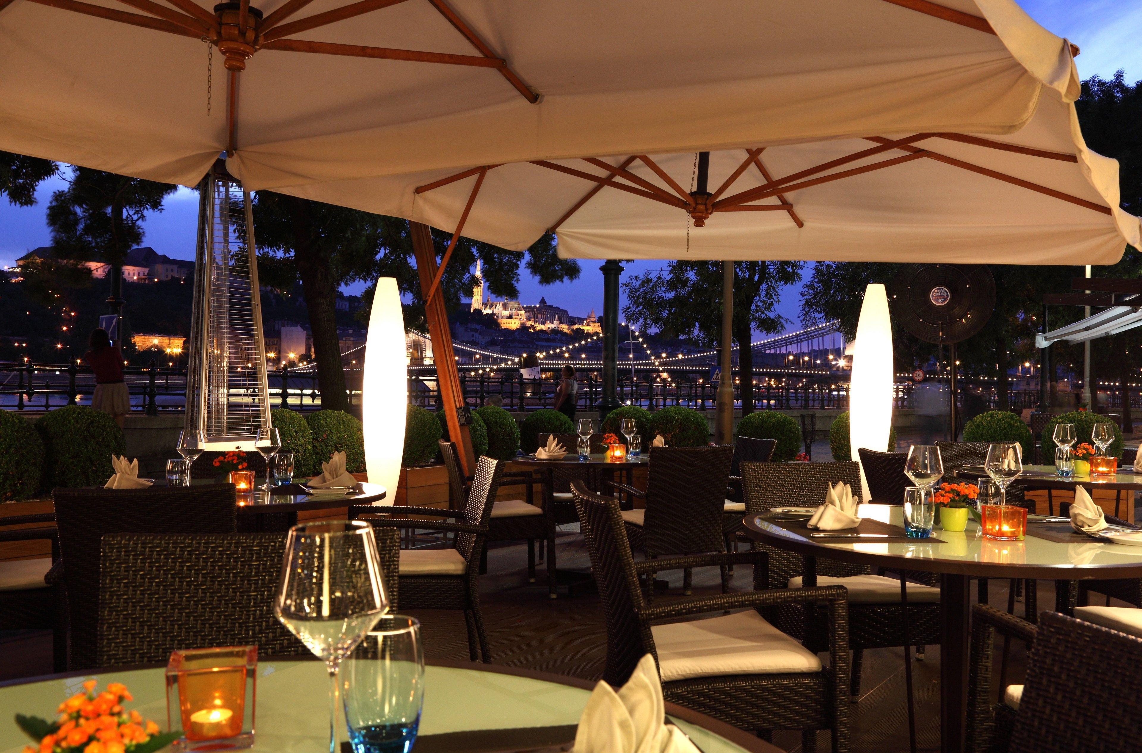 Intercontinental Budapest By Ihg Esterno foto The photo depicts an outdoor dining area at a restaurant, set up for evening service. There are several tables arranged with elegant place settings, including wine glasses and decorative napkins. The space is illuminated by soft lighting from candles