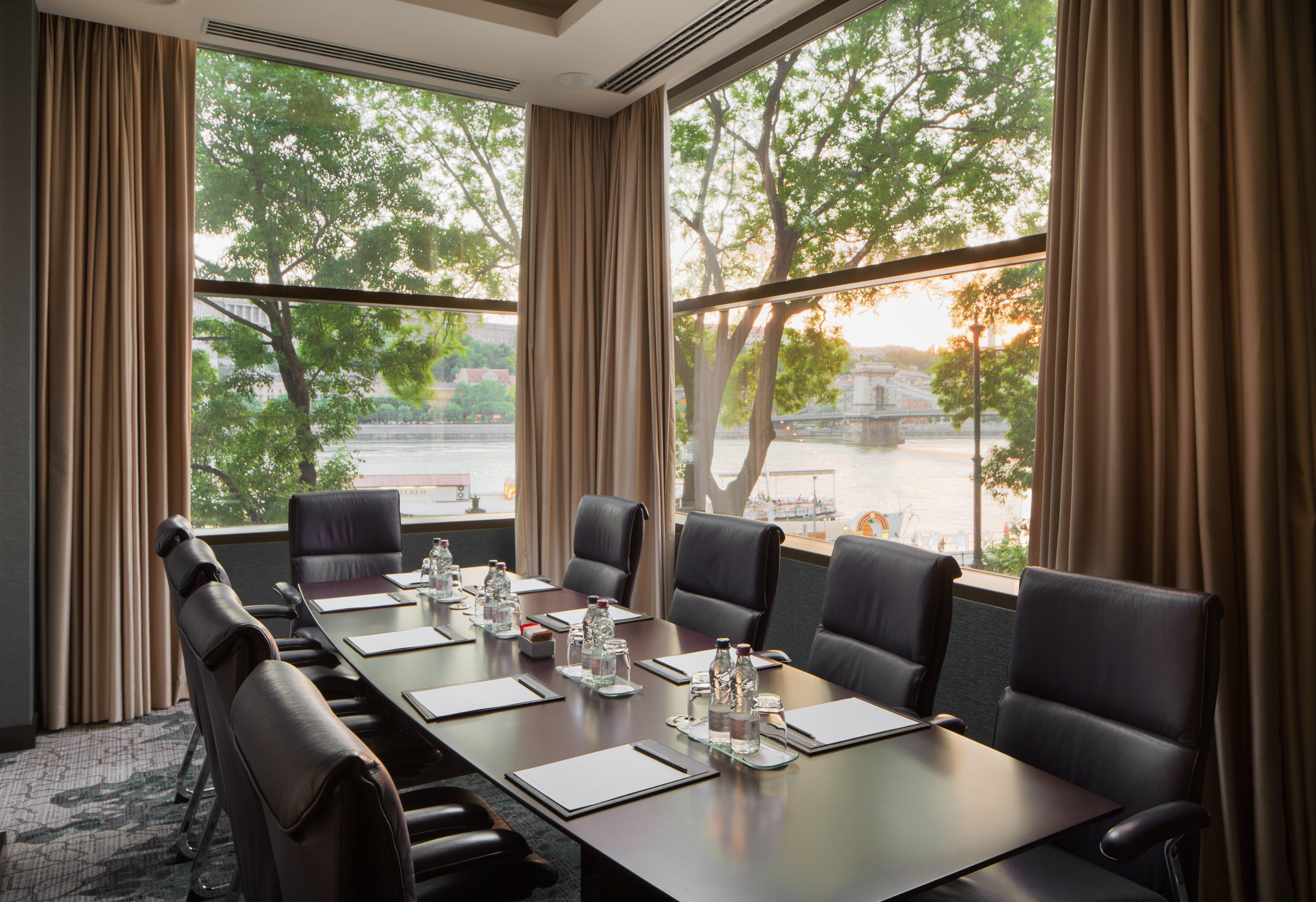 Intercontinental Budapest By Ihg Esterno foto The photo depicts a modern conference room with a long table surrounded by black office chairs. The table is set with glass water pitchers and drinking glasses. Large windows offer a view of greenery outside, with sunlight streaming in and casting a 