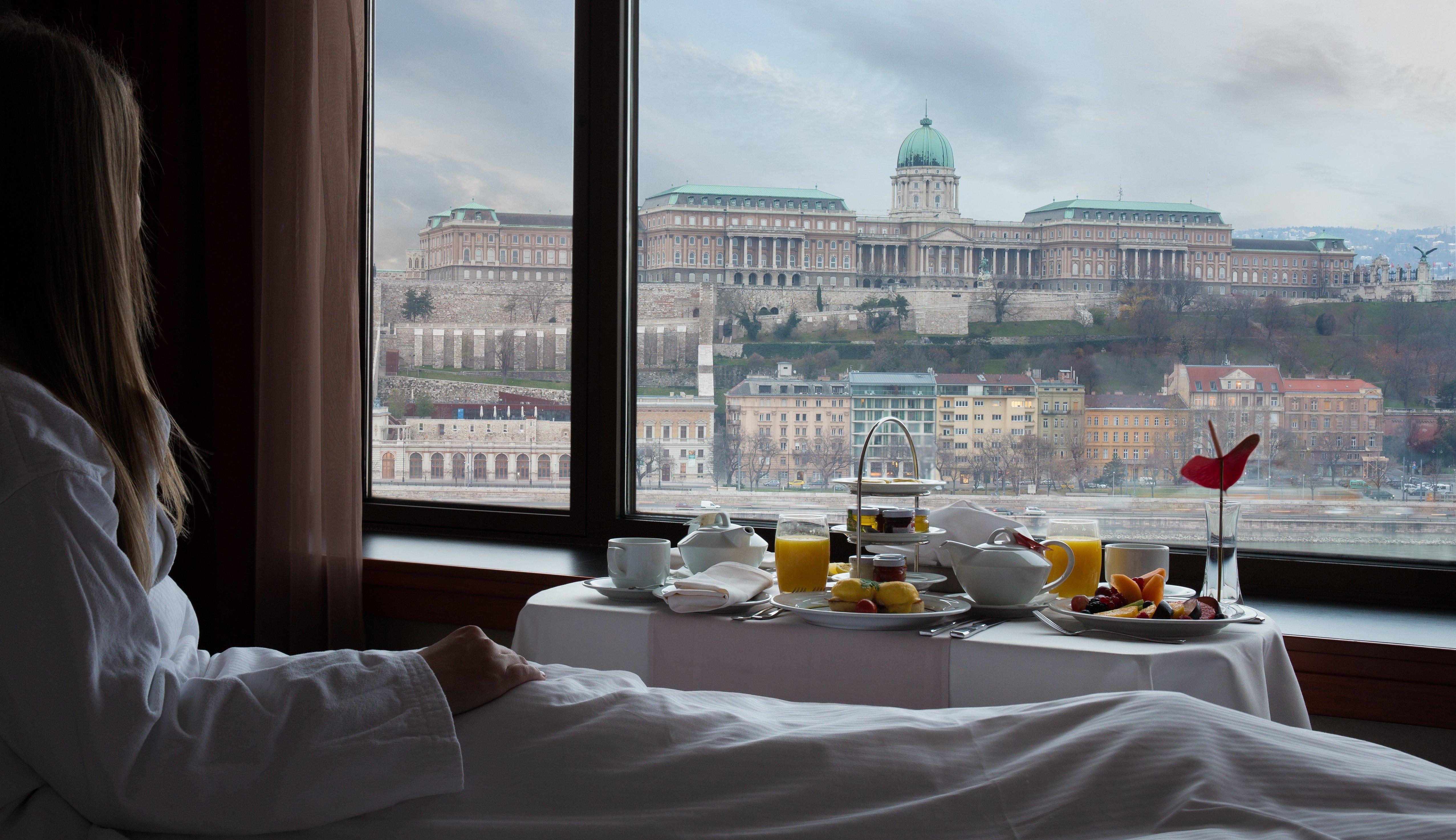 Intercontinental Budapest By Ihg Esterno foto The photo depicts a serene breakfast scene in a hotel room, with a person in a bathrobe reclining on a bed. In the foreground, there is a neatly arranged breakfast spread on a table, including beverages and fruits, set against a backdrop of a beautif