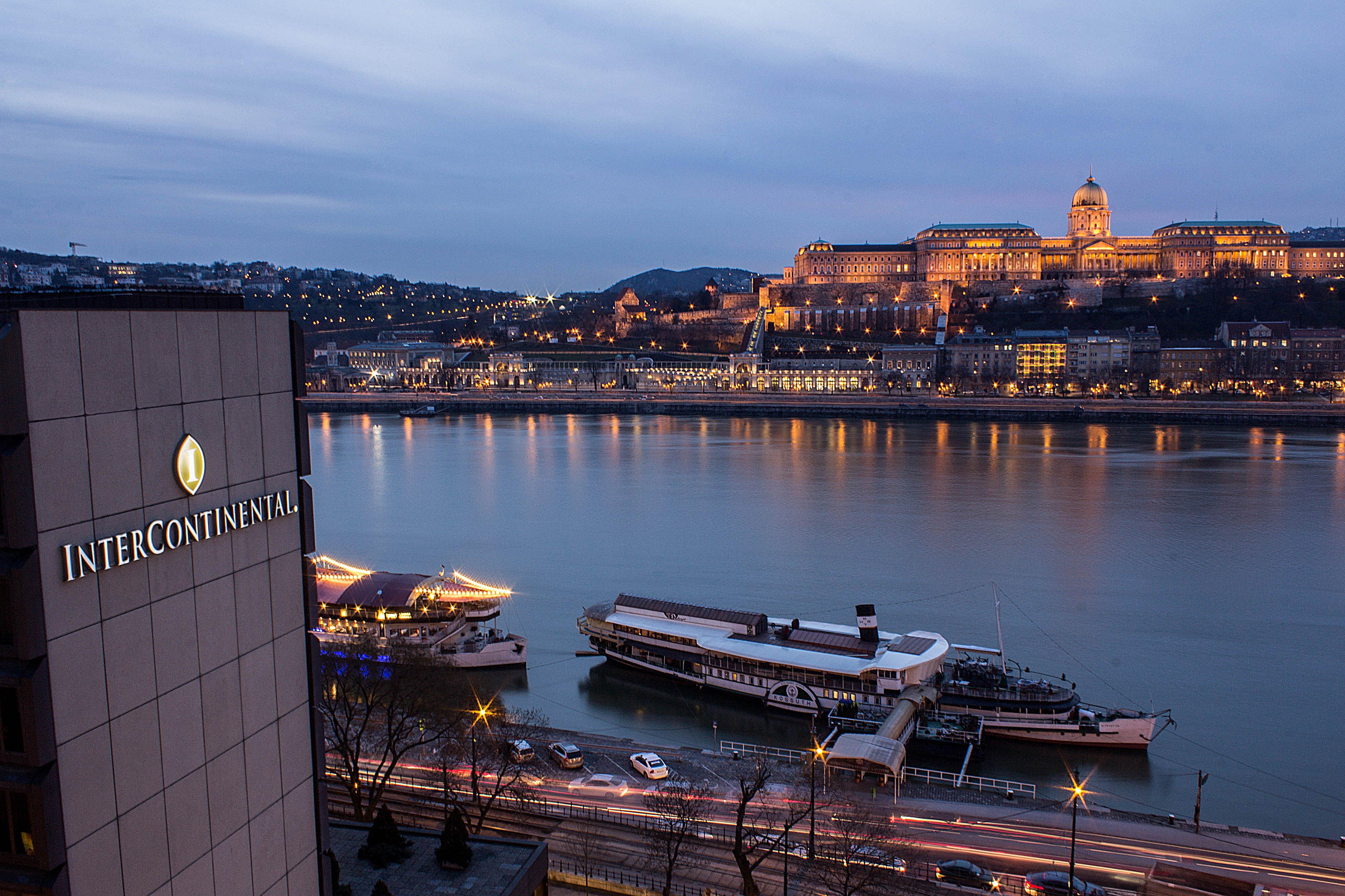 Intercontinental Budapest By Ihg Esterno foto The photo shows a nighttime view of a river, likely the Danube, with buildings illuminated along the shore. In the foreground, there is a hotel, identifiable by its logo, and in the background, a prominent structure atop a hill is lit up, possibly a 