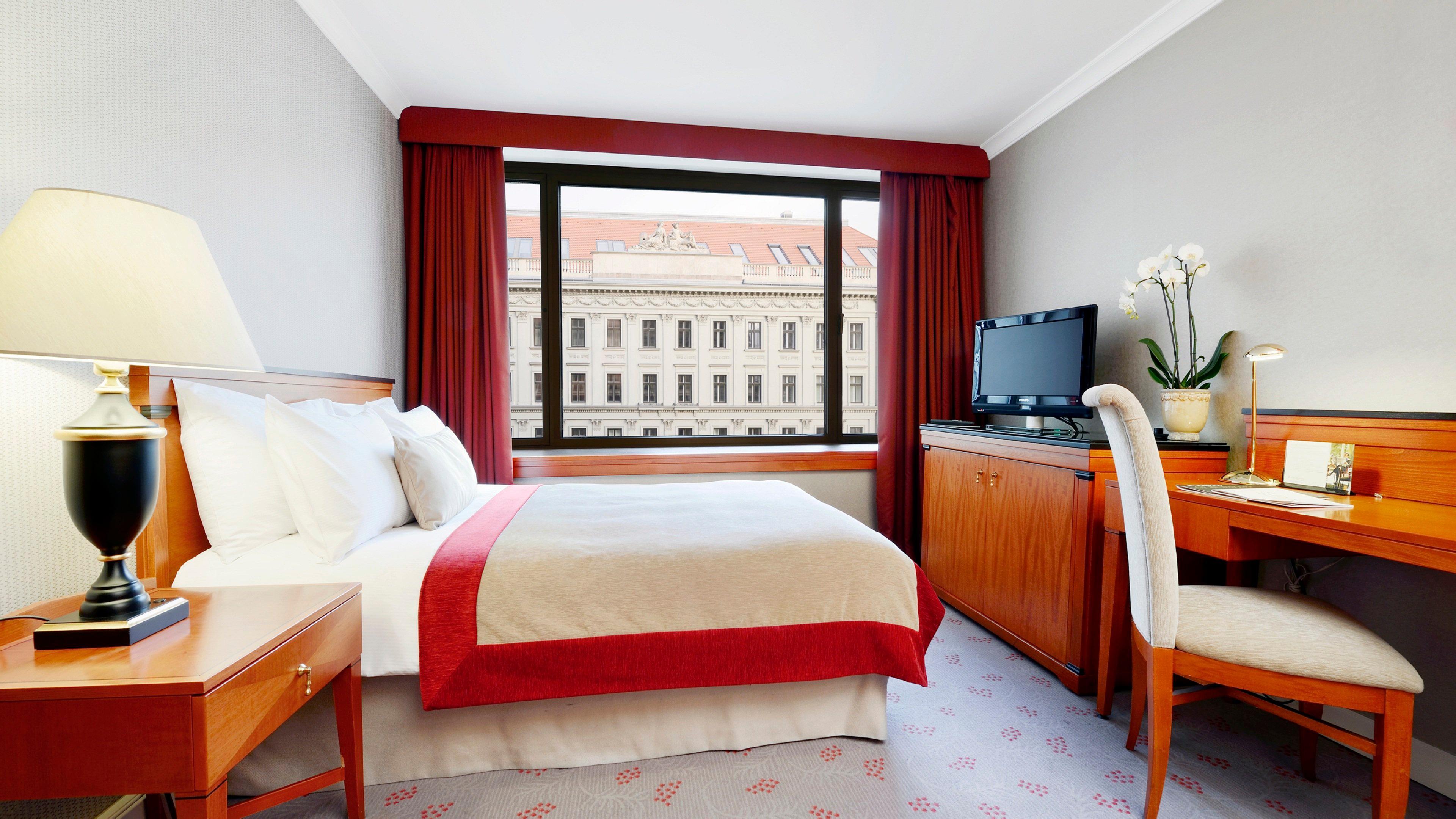 Intercontinental Budapest By Ihg Esterno foto The photo shows a hotel room that features a neatly made bed with white linens and a red accent blanket. There is a large window with red curtains that lets in natural light, revealing views of an adjacent building. A wooden desk and chair are positi