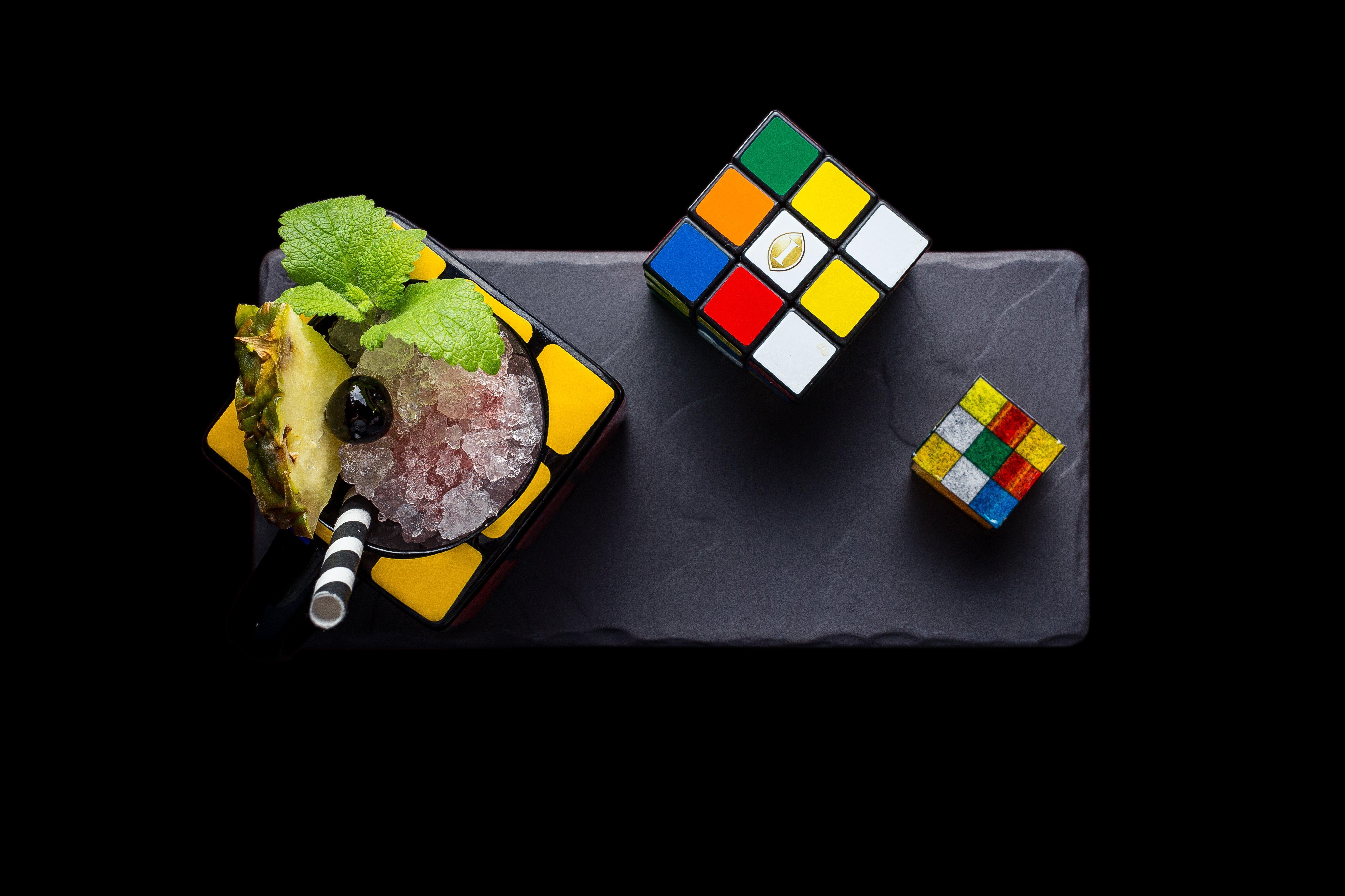 Intercontinental Budapest By Ihg Esterno foto The photo shows a creative arrangement featuring a cocktail or dessert served in a colorful, square dish. The dish is garnished with mint leaves and a slice of pineapple, along with a black olive on top. Next to the dish, there are two Rubik's Cube p