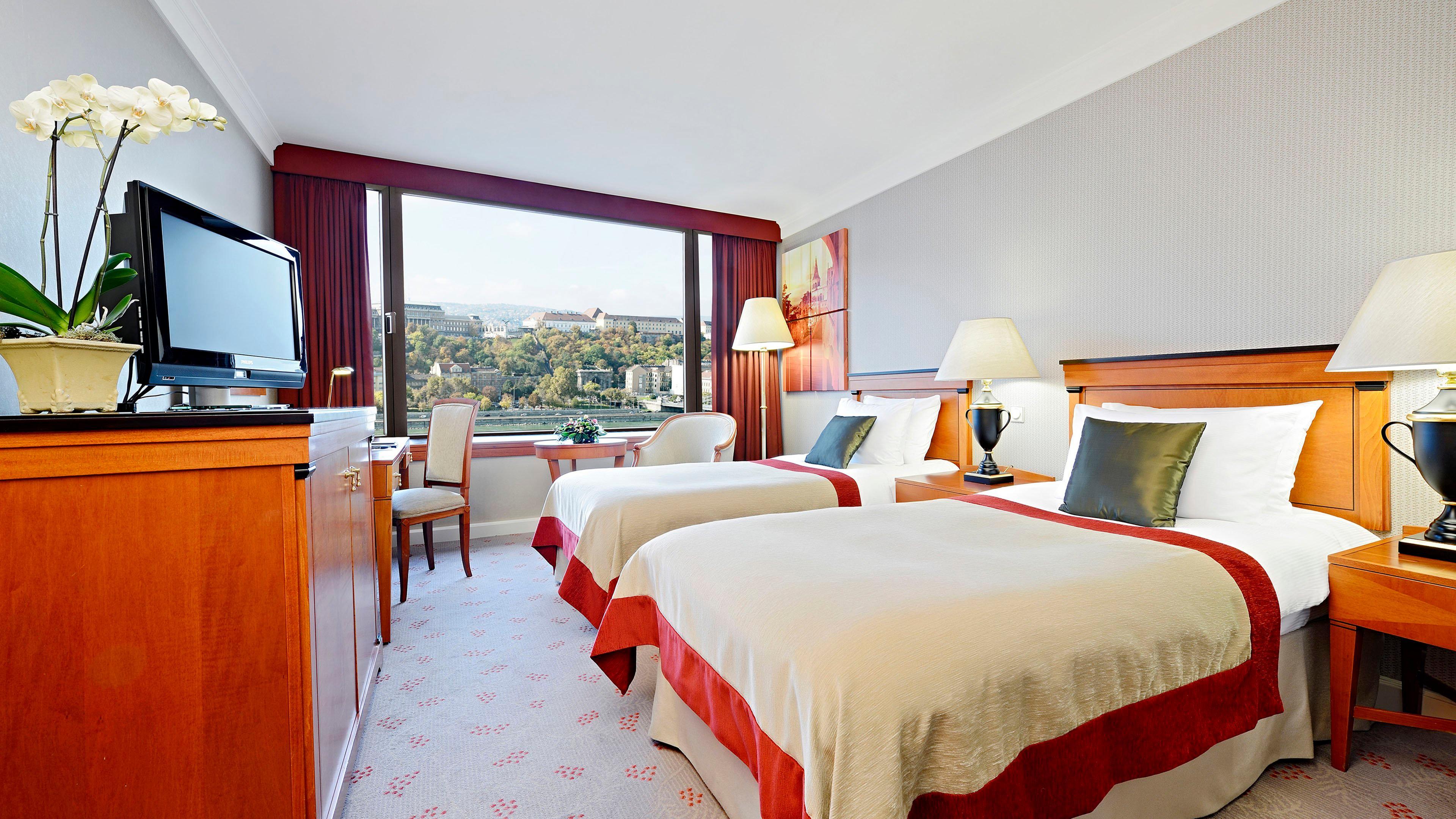 Intercontinental Budapest By Ihg Esterno foto The photo shows a hotel room featuring two twin beds with light-colored bedding and red accents. There is a large window allowing natural light to fill the space, with a view of an outdoor landscape. A wooden dresser and a television are positioned a