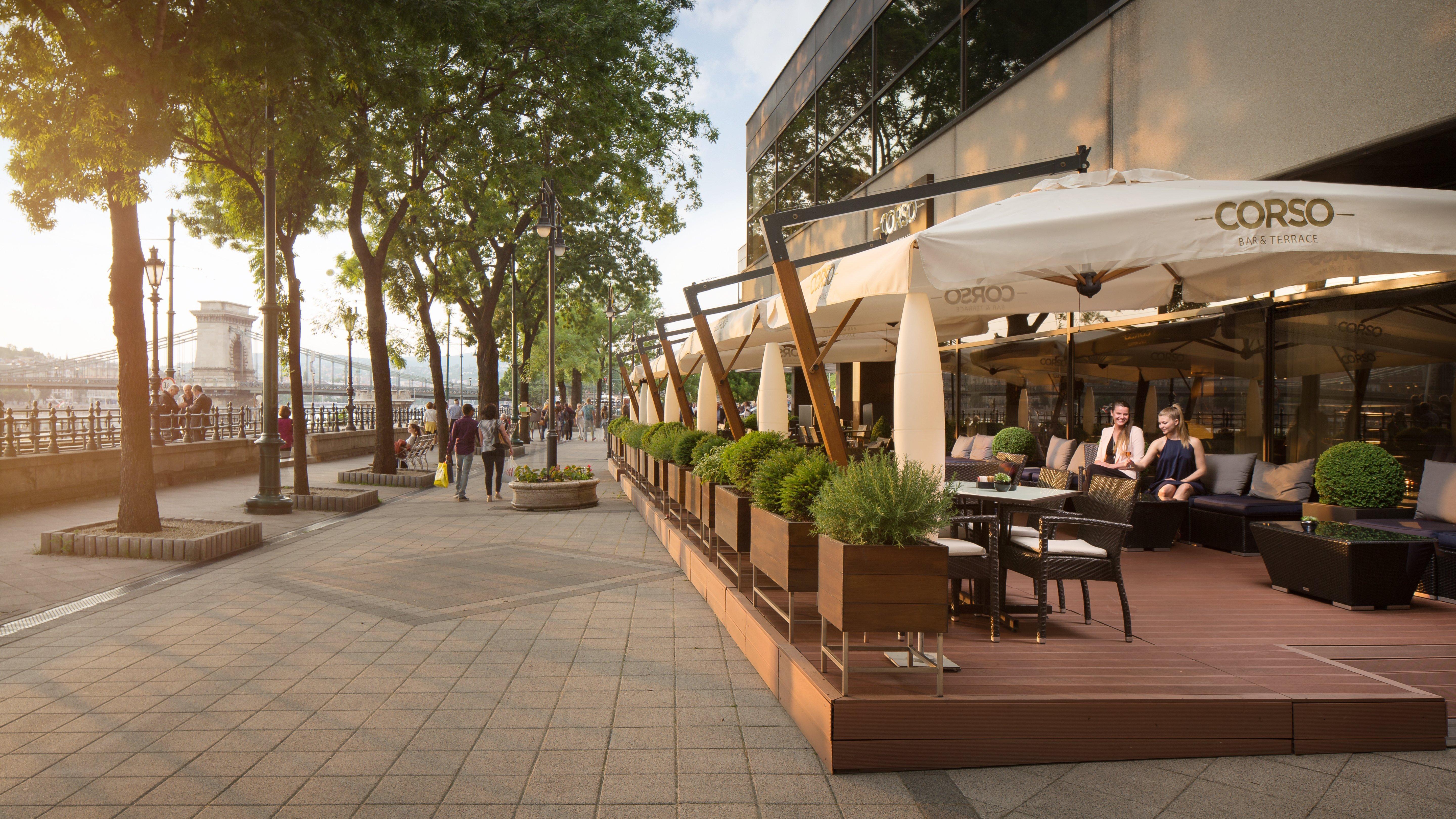 Intercontinental Budapest By Ihg Esterno foto The photo depicts an outdoor seating area of a café or restaurant. It features modern wooden furniture arranged under large, white umbrellas, providing shade for guests. Lush greenery and potted plants are visible along the seating area, enhancing th