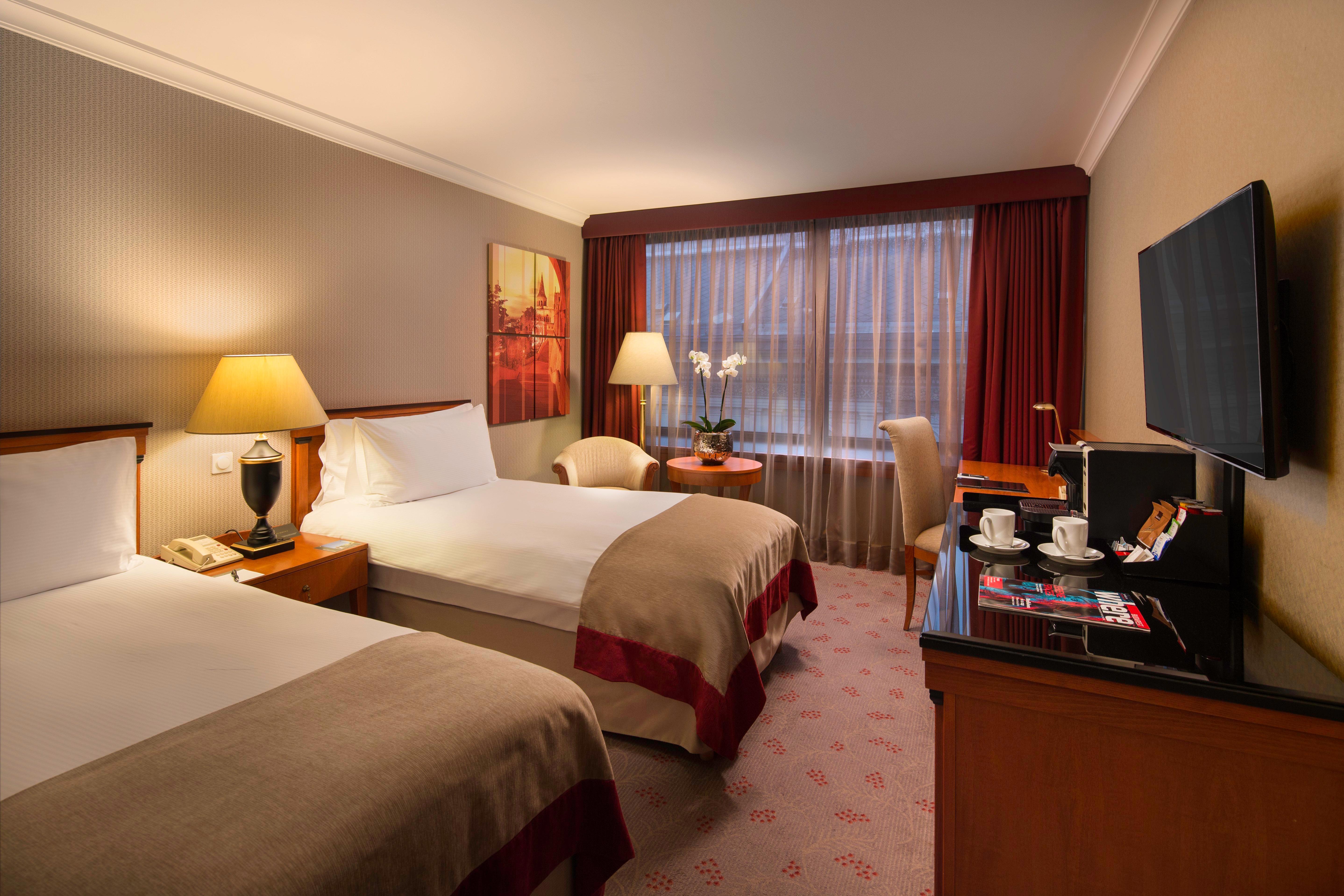 Intercontinental Budapest By Ihg Esterno foto The photo shows a hotel room featuring two twin beds with beige and red bed linens. There are bedside tables with lamps next to each bed. A window with sheer curtains allows natural light to fill the space, and there are two chairs positioned near a 