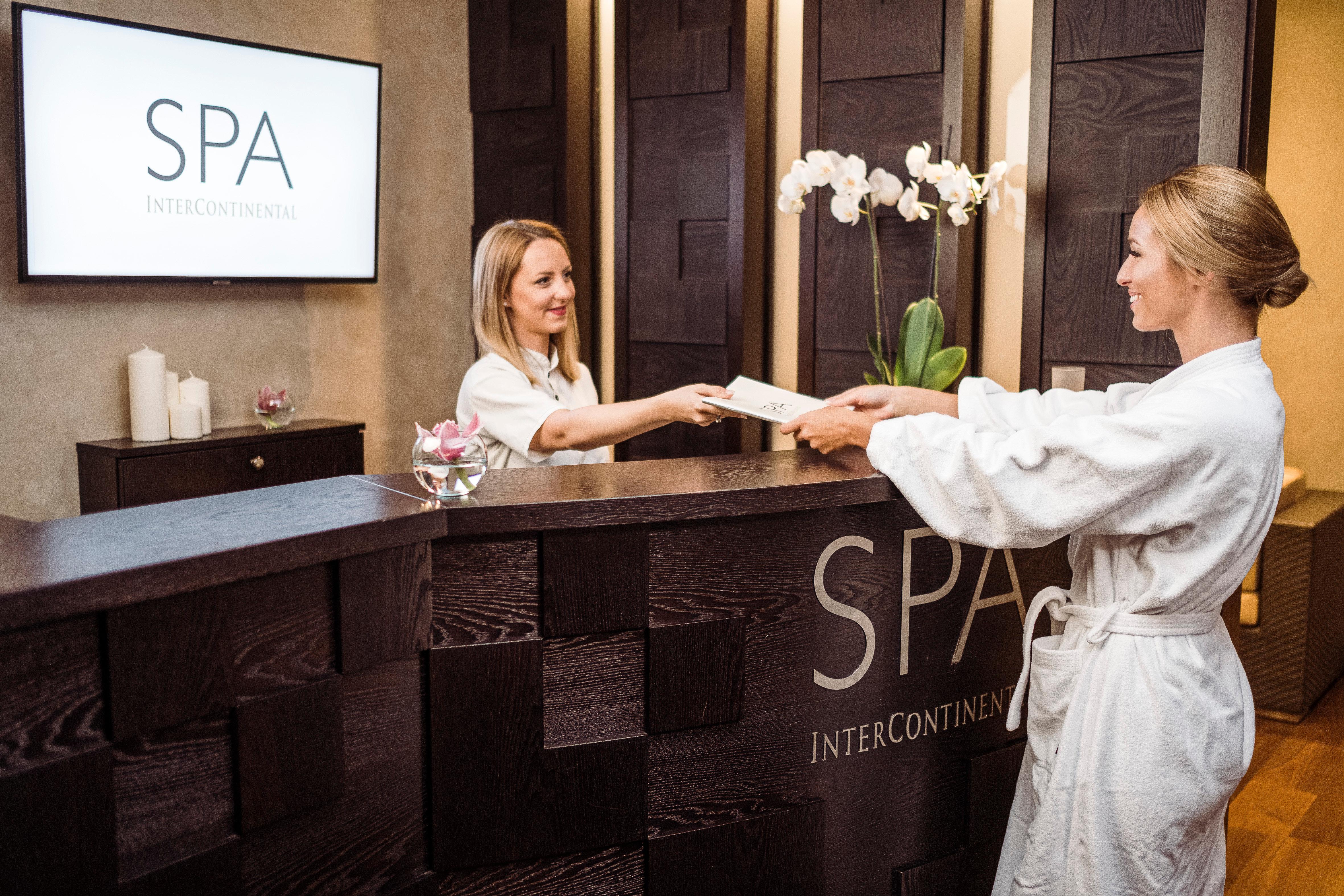 Intercontinental Budapest By Ihg Esterno foto The photo shows a spa reception area. A woman wearing a white robe is handing a document to a receptionist, who is also a woman. The receptionist is standing behind a stylish dark wooden desk that features the word "SPA" prominently displayed. The ba