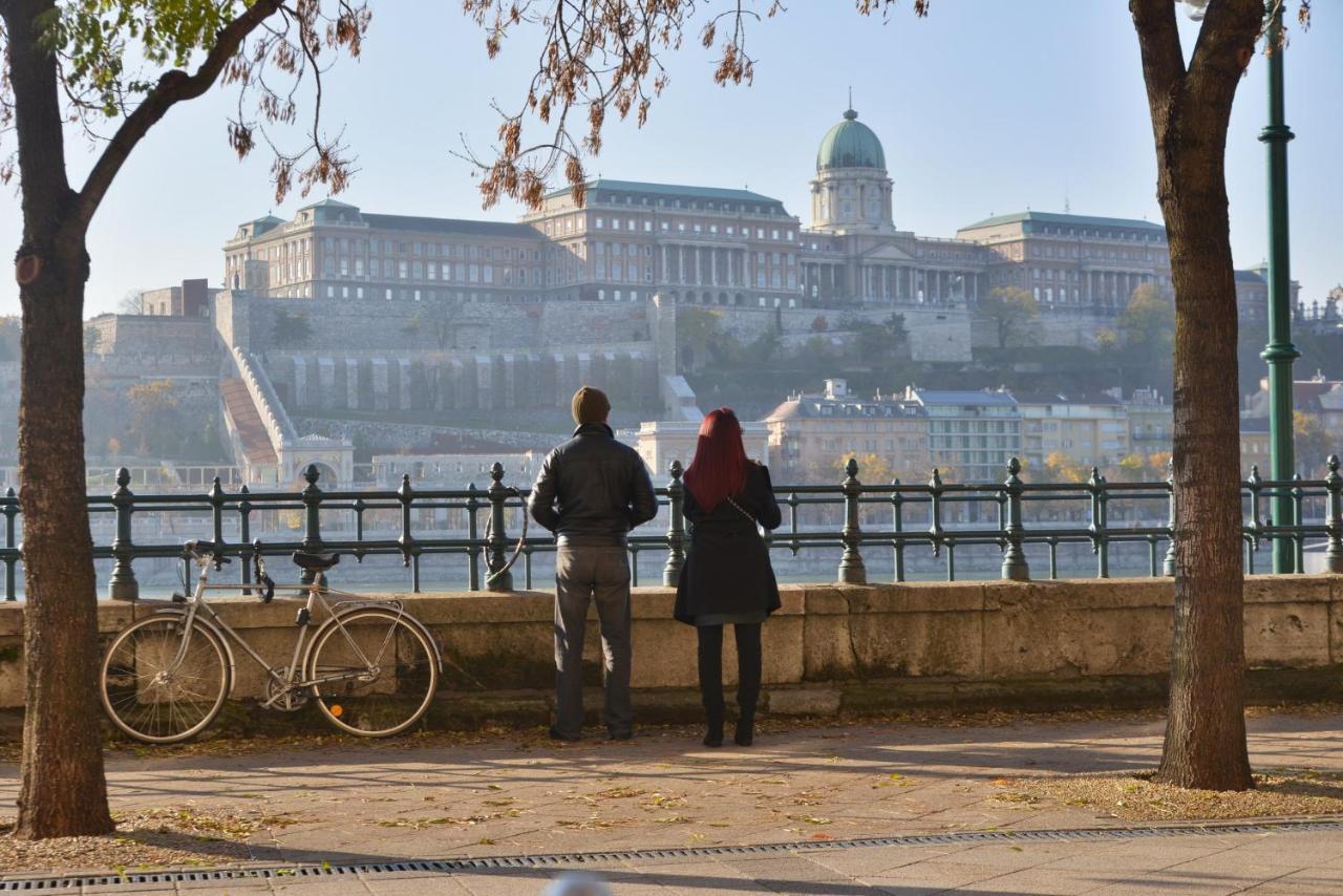 Intercontinental Budapest By Ihg Esterno foto The photo depicts two people standing by a pathway, overlooking a scenic view of a large building complex situated on a hillside, which likely represents a historic site or palace. The individuals are facing away from the camera, with one person wear