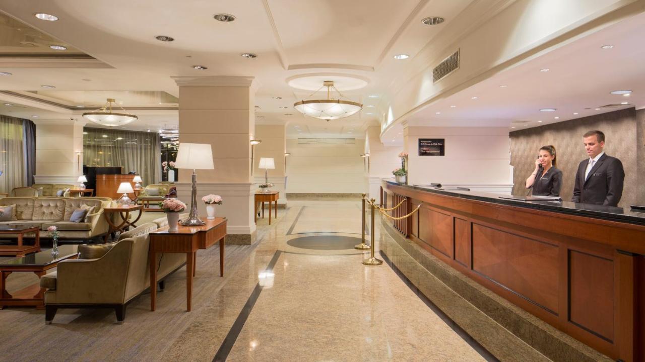 Intercontinental Budapest By Ihg Esterno foto The photo shows a hotel lobby featuring a reception area on the right side. The reception desk is made of dark wood, and there’s a staff member attending to guests. The lobby has elegant decor, with soft lighting from ceiling fixtures and table lamps
