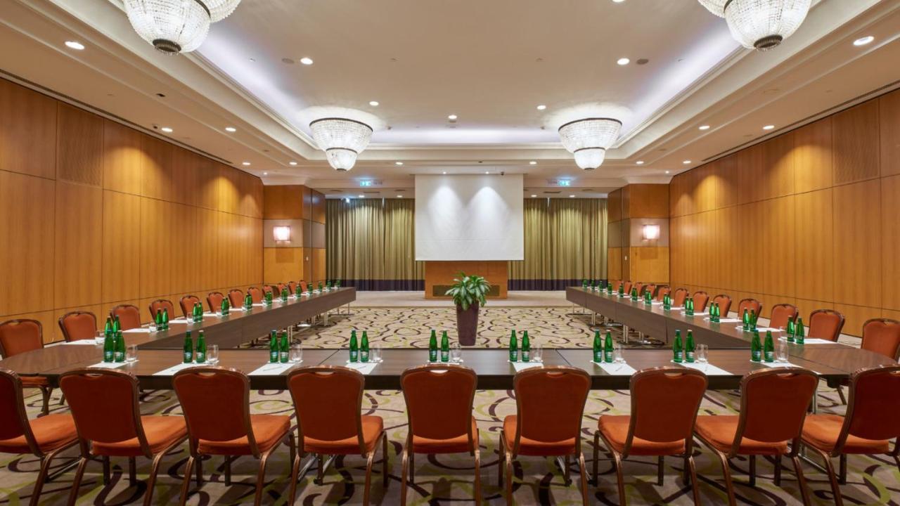 Intercontinental Budapest By Ihg Esterno foto The photo shows a conference room setup. There are several rectangular tables arranged in a U-shape, surrounded by orange chairs. The tables are adorned with glass bottles of water. At the front of the room, there's a projector screen against a backd
